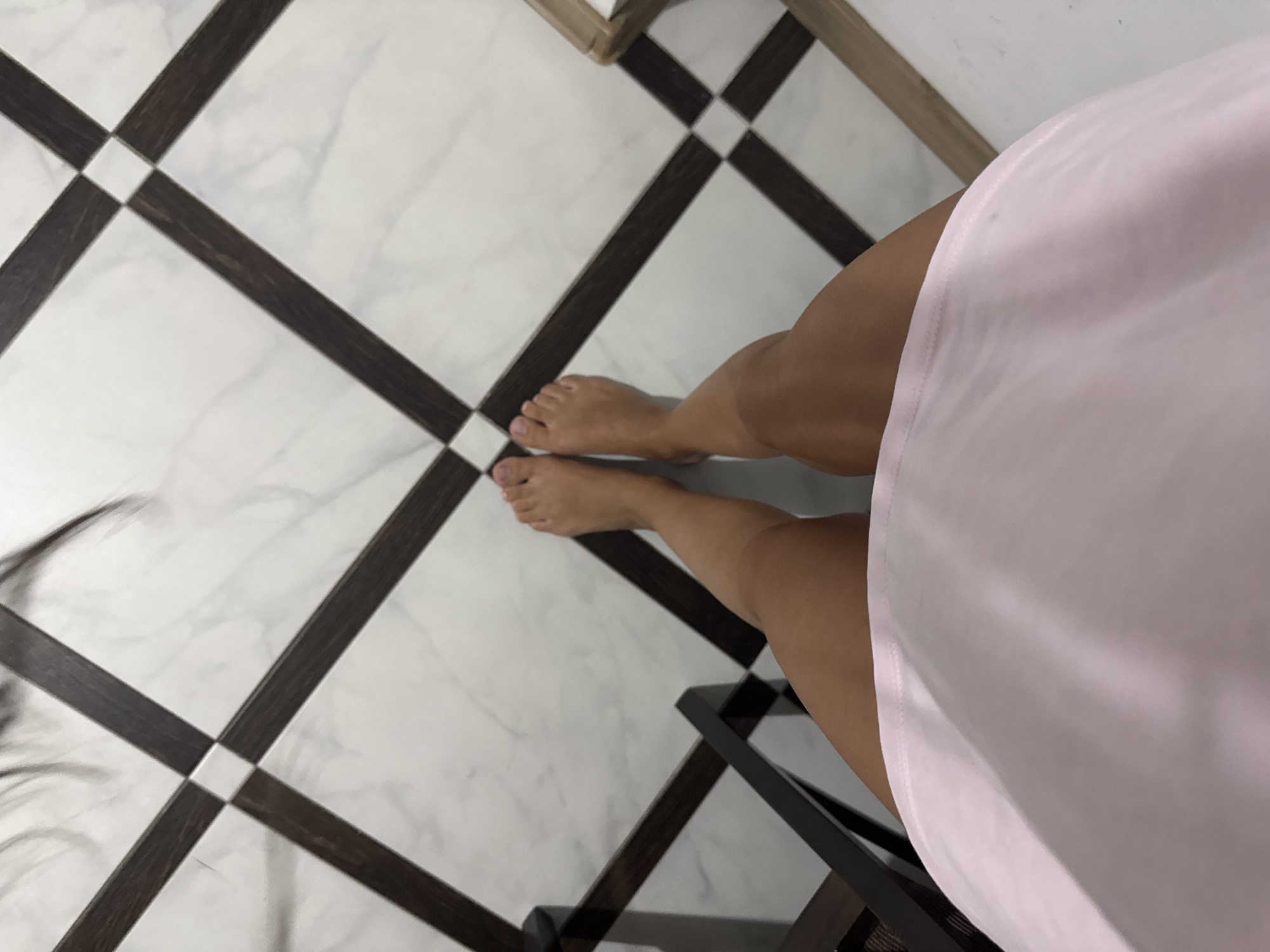 A person's bare feet and legs are visible in the foreground, standing on a marble floor with a dark brown and white tiled pattern. The person is wearing a light pink garment that covers their lower torso and upper legs. The scene appears to be indoors, possibly in a modern home or building. There are wooden furniture elements to the left and right of the feet. No specific landmarks or identifiable features suggest a location within Chișinău, Moldova.