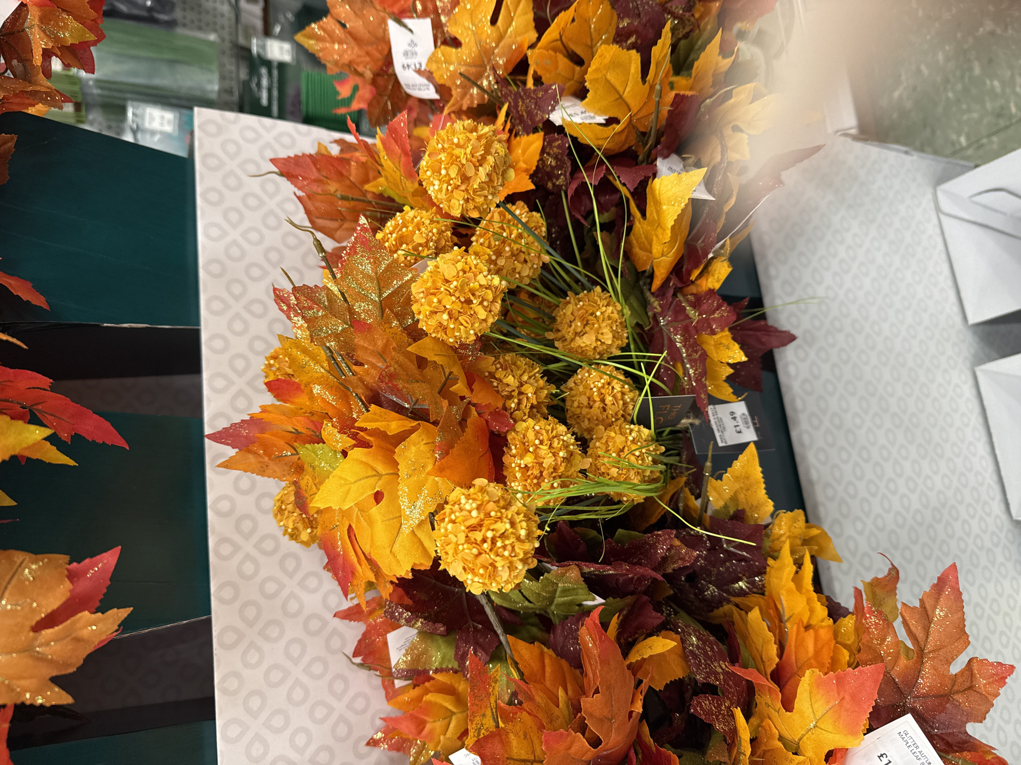 This image captures a close-up view of artificial autumn foliage, likely for decoration, displayed in a retail environment. The arrangement consists of maple leaves in vibrant shades of orange, red, and yellow, many of which are dusted with gold glitter, giving them a shimmering effect. Interspersed among the leaves are small, round, yellow floral orbs, possibly artificial flowers, with tiny white bead-like accents. The foliage is densely packed, creating a rich and textured display.

A white price tag, partially visible and showing "£1.49", is attached to some of the foliage, indicating it is a product for sale. Another tag, also partially visible, seems to be a product description, with text like "Glitter Autumn Maple Leaf". The display sits on a white surface with a subtle, repeating circular pattern. In the background, there is a glimpse of a dark teal surface and a shelf containing various items, some of which are in green packaging.

The lighting suggests an indoor setting, possibly a store aisle or display area. The overall impression is one of seasonal decoration, evoking an autumnal theme. While the image provides a detailed view of the product, it does not offer any specific clues about its precise location within the city of Darlington, United Kingdom, beyond the implication of a retail store setting.