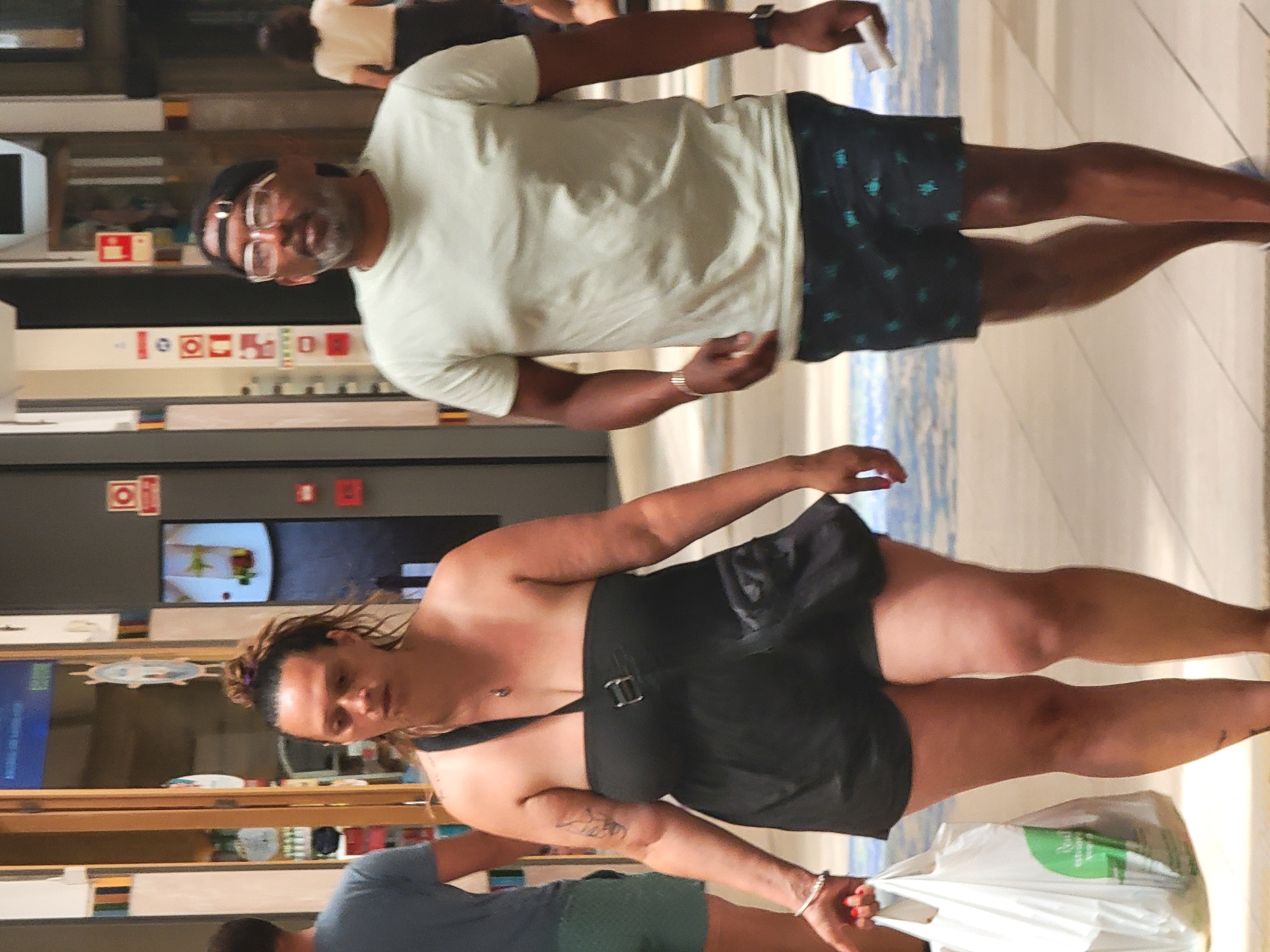 Two individuals are walking through an indoor shopping center. The person on the left, a woman with her hair pulled up in a bun, is wearing a strapless black dress and carrying a white shopping bag. She has a tattoo on her left arm and is wearing a silver bracelet. The person on the right, a man wearing glasses and a black baseball cap, is dressed in a light-colored t-shirt and patterned shorts. He is wearing a watch on his left wrist. The background suggests the environment is a commercial area with storefronts and signs. The lighting is artificial and appears to be typical for an indoor retail space. This scene could be located in the city of Faro, Portugal, within one of its shopping centers.