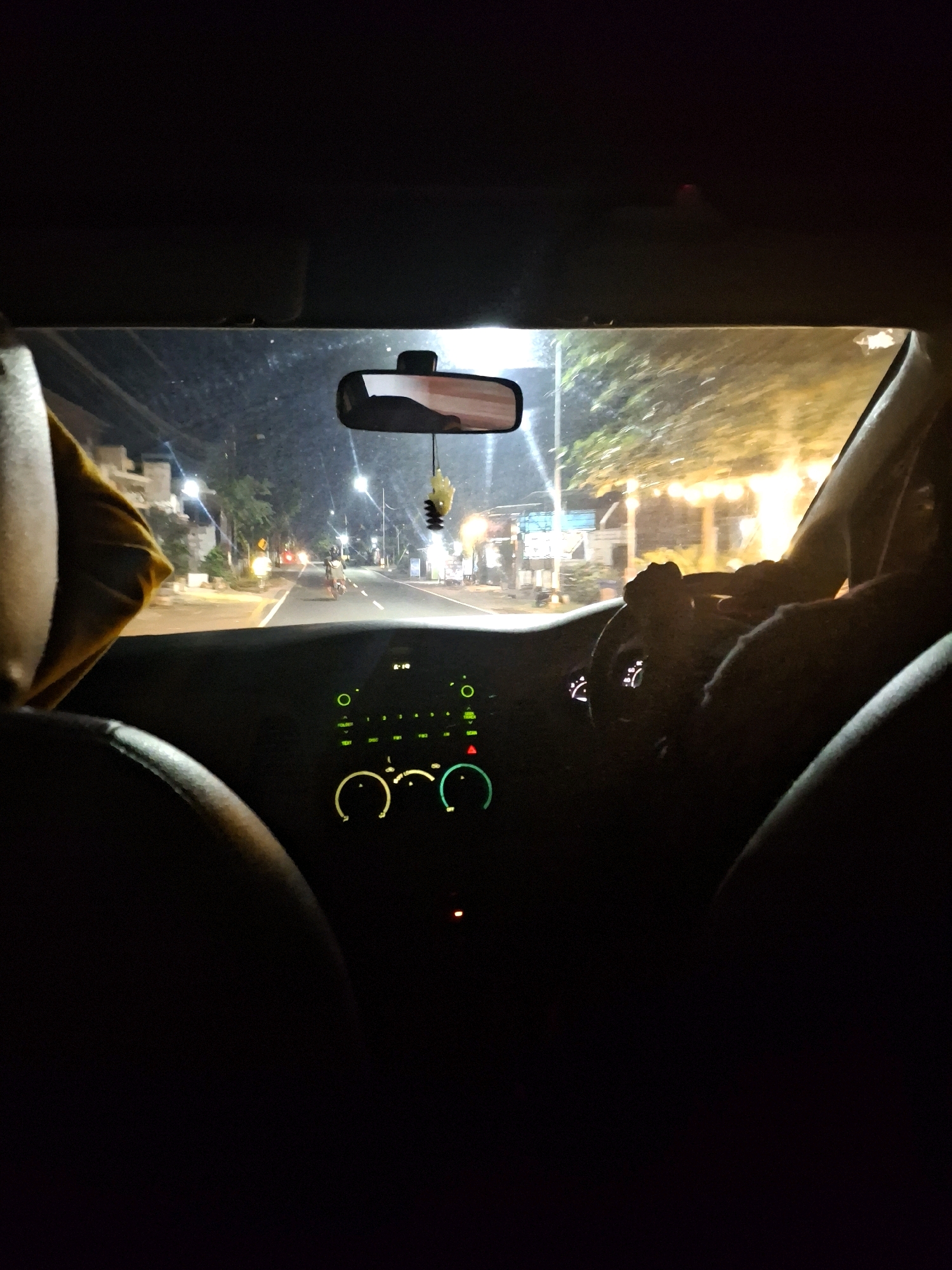 The image, taken from inside a vehicle at night in Lumajang, Indonesia, shows a view through the windshield. The road ahead is illuminated by streetlights and the headlights of oncoming traffic. On the right side of the road, there are buildings and trees, some adorned with decorative lights. A person is seen riding a motorcycle in the distance, heading away from the camera. Inside the car, the dashboard is visible, with glowing green and white indicators. The steering wheel and a person's hands gripping it are also present, indicating the vehicle is in motion. The overall scene suggests a typical night drive through a populated area of Lumajang.