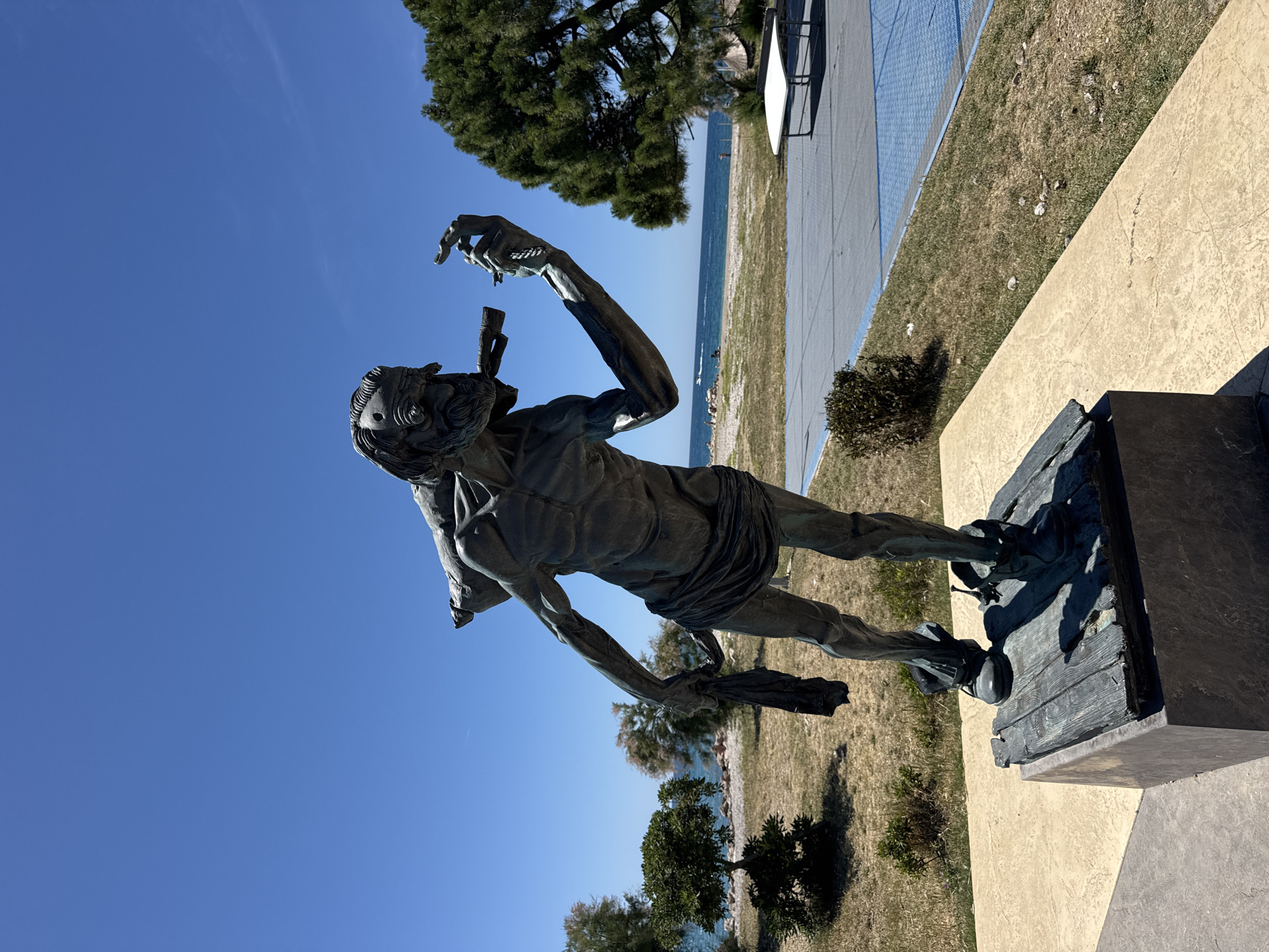 A bronze statue of a blindfolded man is located outdoors in the city of Bar, Montenegro. The statue is placed on a dark bronze, wood-looking platform, which is seated on top of a light brown concrete pedestal. The man is standing and has a torn piece of cloth in his left hand, and is gesturing with his right hand. The background includes the sky, a body of water, trees, and what looks like a walkway of some sort. The sky is clear and blue. The statue is a weathered bronze color.
