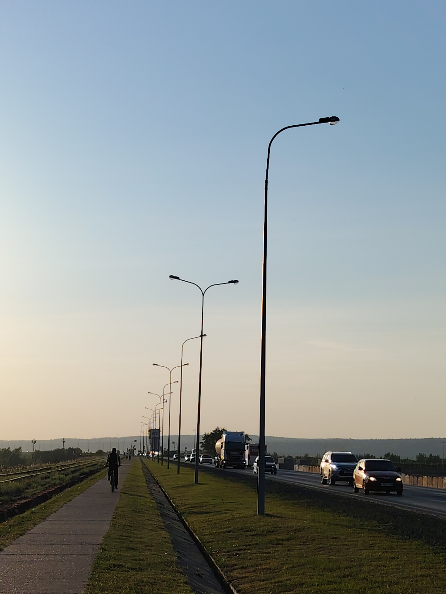 The image shows a road in Chaikovsky, Russia. A person on a bicycle travels along a paved path beside the road. A line of evenly spaced street lamps runs between the path and the road, illuminating the scene. The road is filled with moving traffic consisting of cars and trucks. A strip of green grass separates the path from the road. Beyond the path, there is a railway track. The background shows a line of distant hills. The sky is a pale blue.