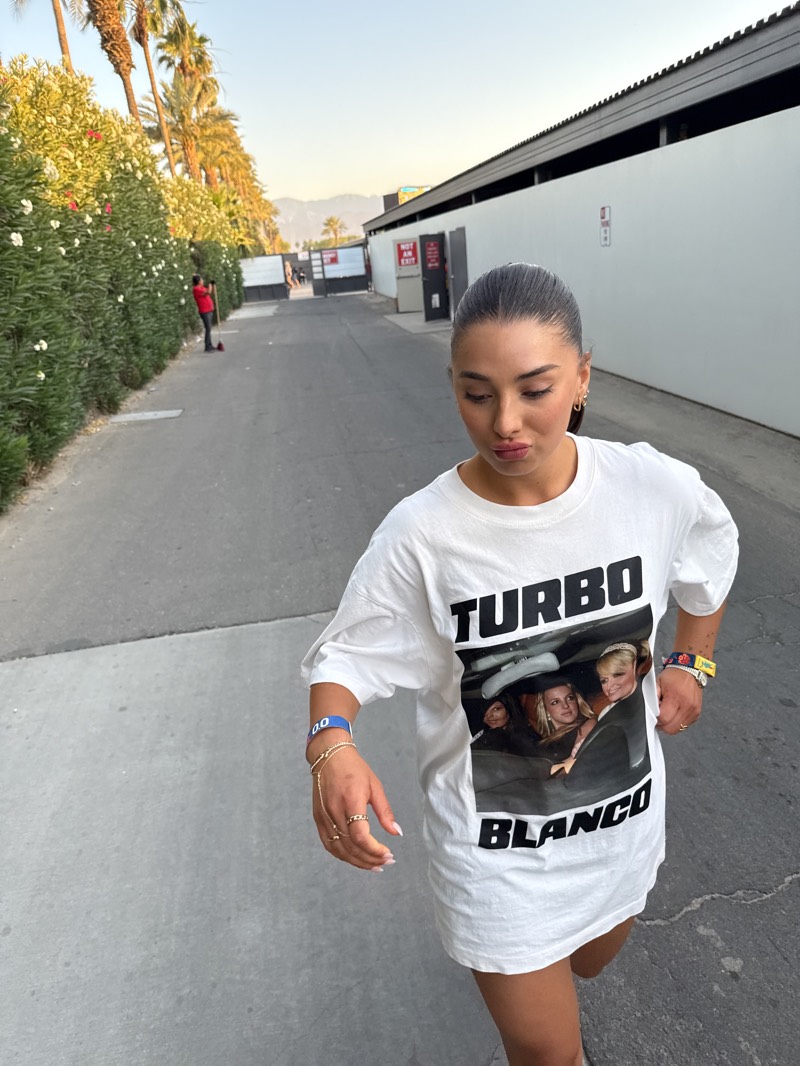 A young woman stands on a paved road in Indio, California, wearing a white T-shirt with the words "TURBO BLANCO" and a picture of three women in a car printed on it. She has her hair pulled back and is wearing jewelry on her wrists and fingers. Behind her, to the left, is a tall hedge with flowering plants, and to the right is a long, white building. In the distance, there are palm trees, mountains, and a gate with signs on either side of a red box. A person in a red shirt is visible near the hedge. The lighting indicates it is either early morning or late afternoon.