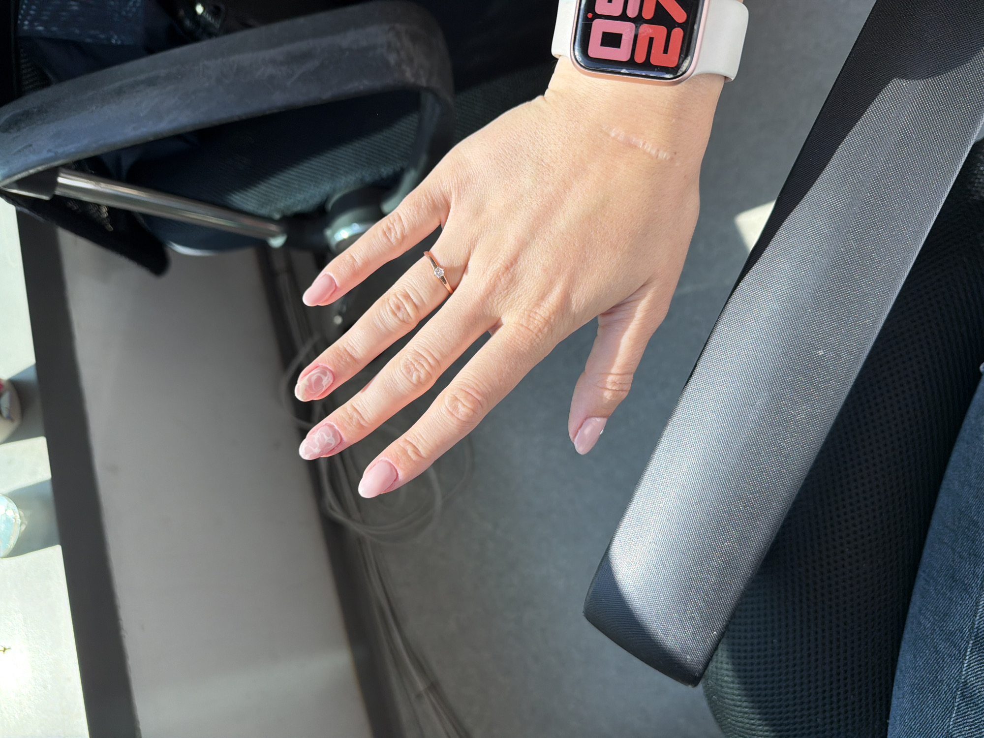 A woman's left hand is shown from a high angle, resting on a dark grey, textured surface. The woman's hand is positioned centrally in the frame, with her fingers extended and slightly curled. Her fingernails are painted a pale pink, with a subtle marble-like design on some of the nails. She is wearing a delicate rose gold ring with a small diamond on her left ring finger. A white Apple Watch with a pink bezel is visible on her left wrist. A visible scar runs horizontally across her inner wrist. The background is out of focus but appears to be an indoor environment with a bright window on the left, casting shadows. The scene does not provide any information to determine the location in Sofia, Bulgaria.