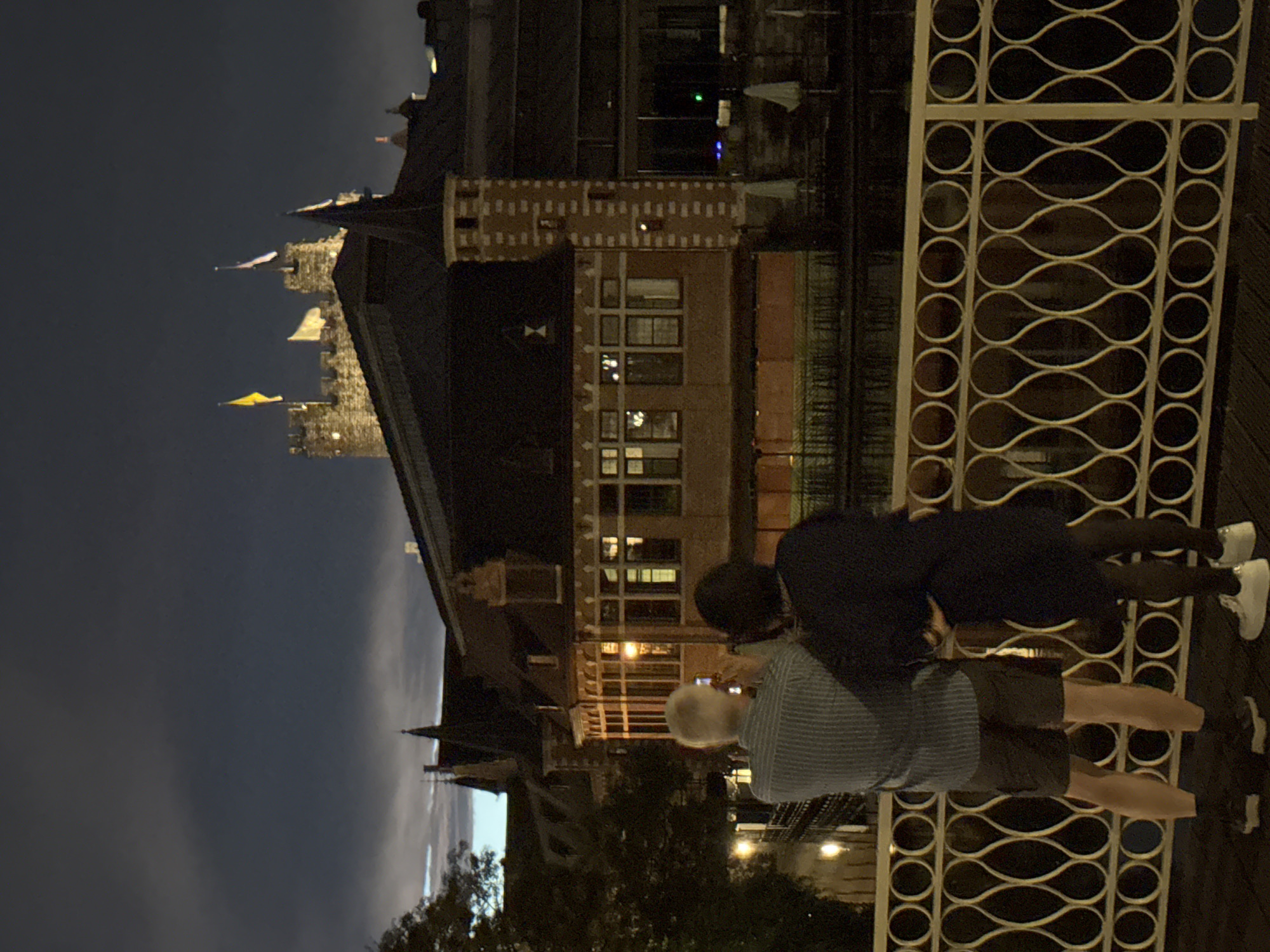 Two individuals, a man and a woman, stand on a bridge at night, facing a canal and buildings. The man, in a striped shirt and shorts, holds a phone, appearing to take a photograph. The woman, in a dark dress and tights, stands beside him, her arm around his shoulder. Behind them, illuminated by internal lights, is a building with large windows and a prominent turret. In the distance, a castle-like structure with flags is visible against a dark sky. The scene is located in Ghent, Belgium, likely near the Gravensteen castle and the canals of the city.