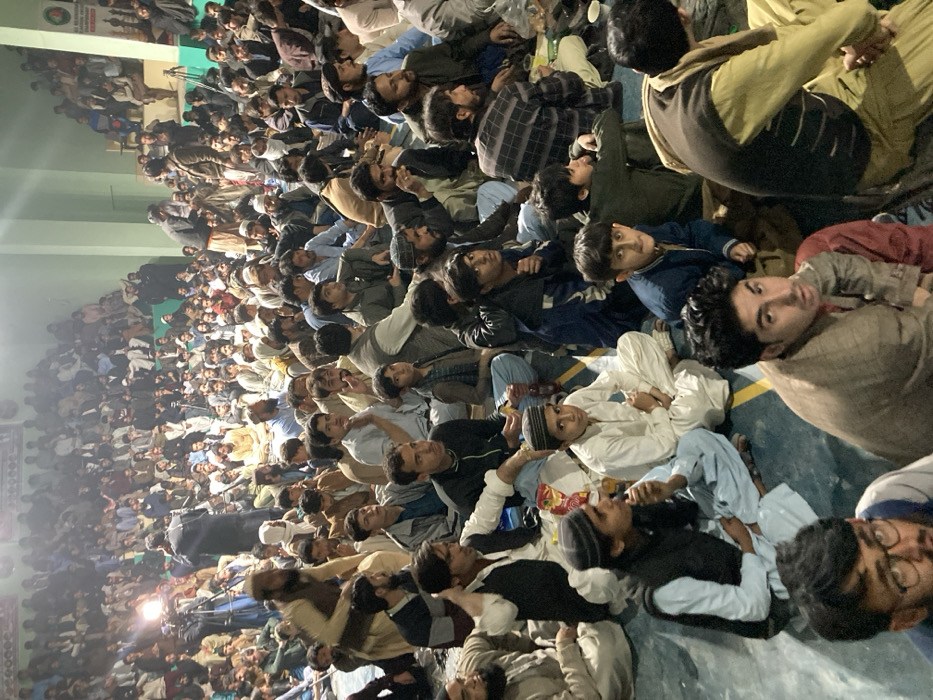 An indoor scene in Quetta City Tehsil, Pakistan, depicts a densely packed male gathering within a large hall. Hundreds of men and boys, predominantly in traditional South Asian attire such as Shalwar Kameez, are seated on the dark-colored floor, which features light-colored court lines. In the mid-ground, a professional video camera on a tripod, equipped with a bright attached light, is positioned to film an event. Two individuals operate the camera setup, one standing directly behind it, and another person holding a microphone is nearby. The majority of the seated audience faces forward, indicating attention towards a central point not fully visible. Some individuals in the foreground and mid-ground consume food or drinks from plastic bottles, plastic bags, and foil containers. The hall features pale green walls, and a partial banner with Urdu script is visible on the upper right wall. The overall lighting is diffuse, supplemented by the bright camera light.