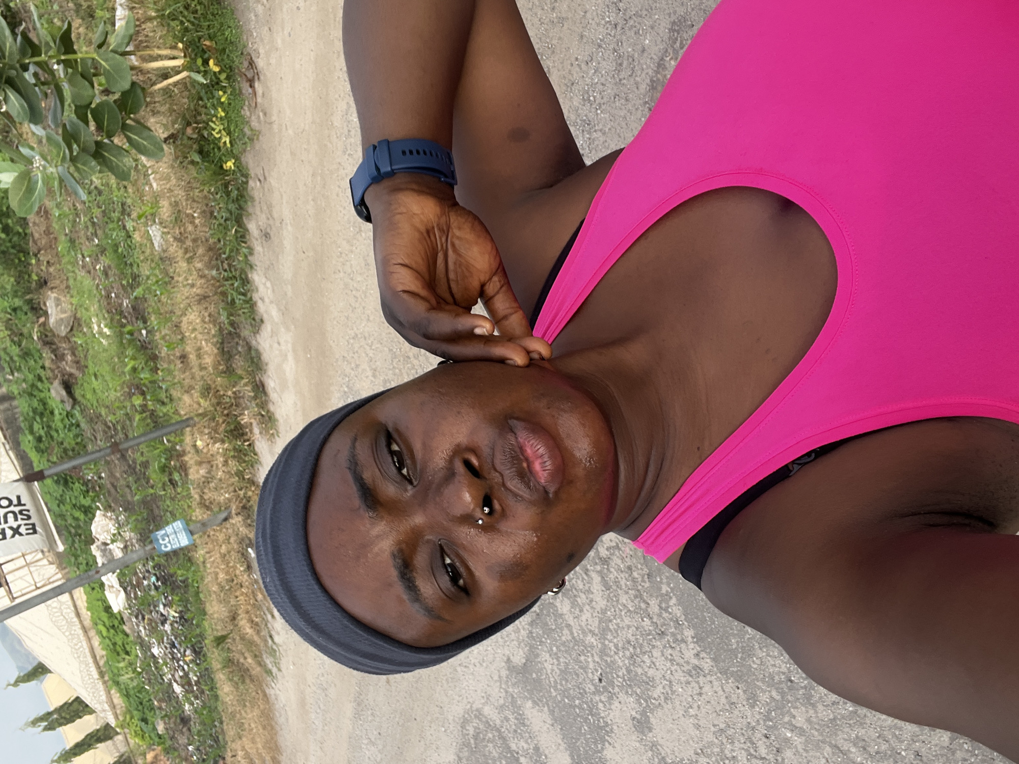 A medium shot taken outdoors shows a Black woman in a bright pink tank top and a dark gray headband. She is looking at the camera with her lips puckered. She has a nose ring and is wearing a dark blue smartwatch on her left wrist. Her right hand is resting on her cheek. The background is a dirt road with sparse vegetation and some trash on the left side. A white sign with black text is visible in the upper left quadrant of the image. The scene appears to be in an undeveloped area, possibly on the outskirts of Abuja, Nigeria.