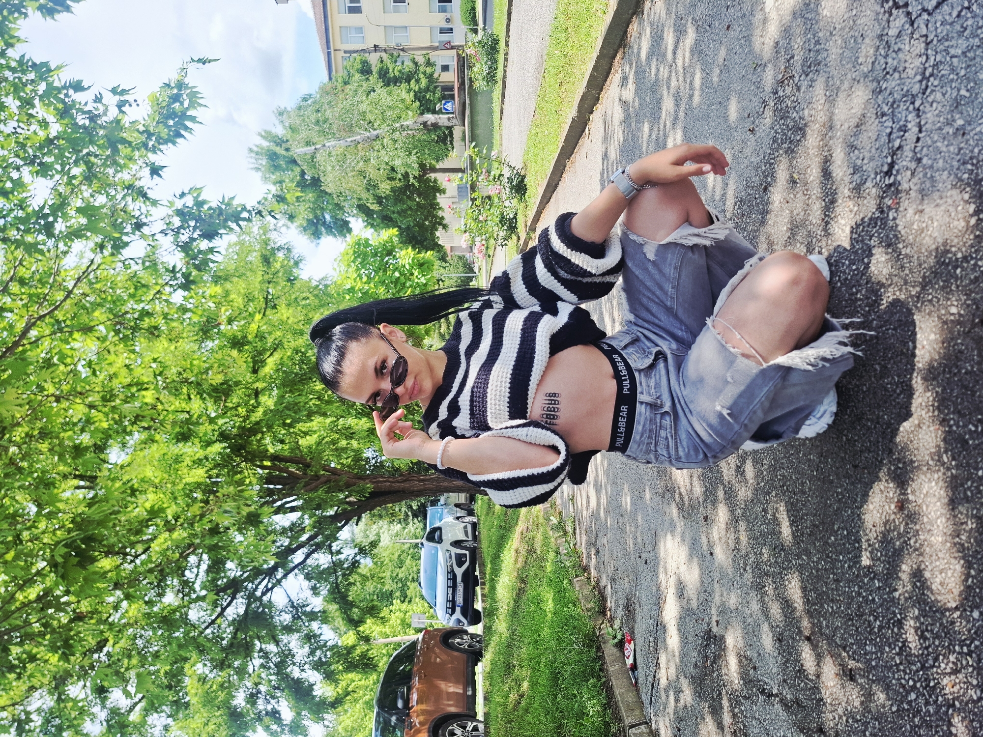 A young woman is captured in a photo, originally rotated 90 degrees clockwise, sitting on the ground outdoors. She has dark hair styled in a high ponytail and is wearing sunglasses. Her expression suggests confidence as she poses for the camera, her right hand adjusting her sunglasses. She is dressed in a black and white striped long-sleeved cropped top and grey ripped denim shorts or jeans. The black elastic waistband of her bottoms, clearly visible, features the text "PULL&BEAR." On her midriff, stylized text, possibly a temporary tattoo, is visible, though partially obscured. She accessorizes with a silver-toned watch or bracelet on her left wrist and a white beaded bracelet on her right.
The setting appears to be a sunny day in Mezdra, Bulgaria, indicated by the clear bright light and sharp shadows cast by trees on the asphalt or concrete ground where she is sitting. Green foliage from numerous trees lines a grassy verge adjacent to the paved area. In the background, multiple cars are parked, including a brown SUV and a white car with a partially visible Bulgarian license plate ("DB 0337 HX"). Beyond the trees and cars, a multi-story building with windows is visible, suggesting a residential or urban environment. The weather is clear and bright.