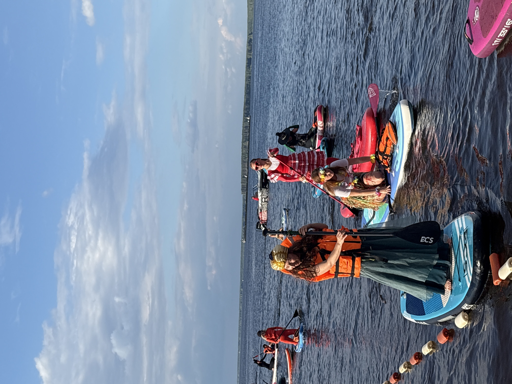 The image depicts a group of people on paddleboards in a body of water, likely a river or lake, under a partly cloudy sky. The location is identified as Chaikovsky, Russia. Several individuals are dressed in costumes, suggesting a themed event. In the foreground, a person wearing an orange life vest and a long green skirt is holding a paddle. To their left, another person in an orange outfit is also on a paddleboard with a paddle. Further back, a person in a red and white dress stands on a paddleboard. A person in a black costume is seen on a paddleboard behind the person in the red dress. A person wearing a lei and a white t-shirt is kneeling on a paddleboard in the mid-ground. Several other paddleboards and individuals are visible in the background, along with a distant shoreline with trees. The water is rippled, indicating some movement. A string of buoys is present in the foreground, marking a boundary or path.