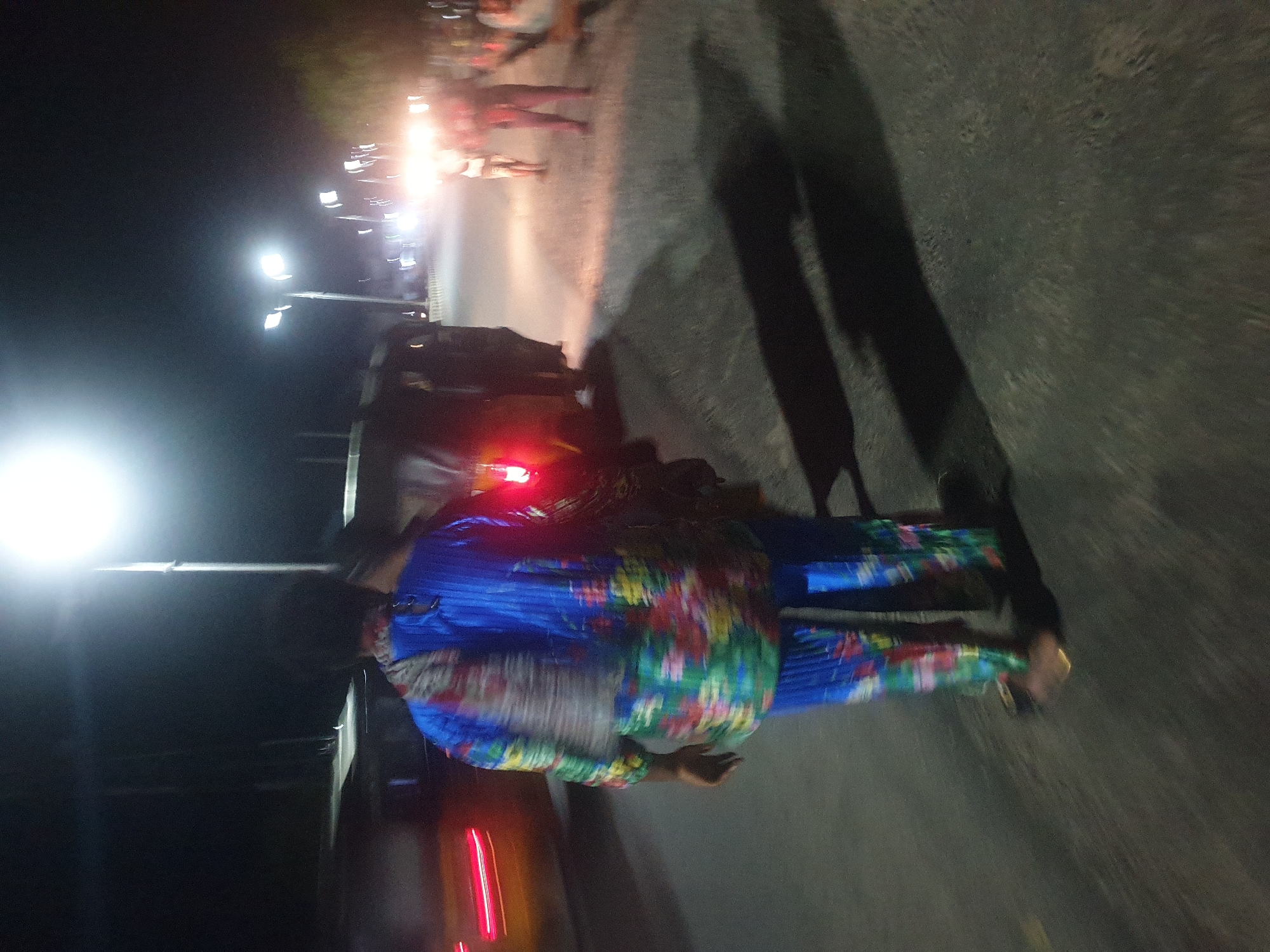 At night in Zango-ndaji, Nigeria, a person wearing a blue outfit with a colorful floral pattern is standing on a paved road, perpendicular to the camera's perspective. Street lights illuminate the scene, creating bright points of light in the dark background. A vehicle with red tail lights is visible near the person. Other people are present in the background under the street lights. Long shadows are cast on the road from the subjects and the lighting.