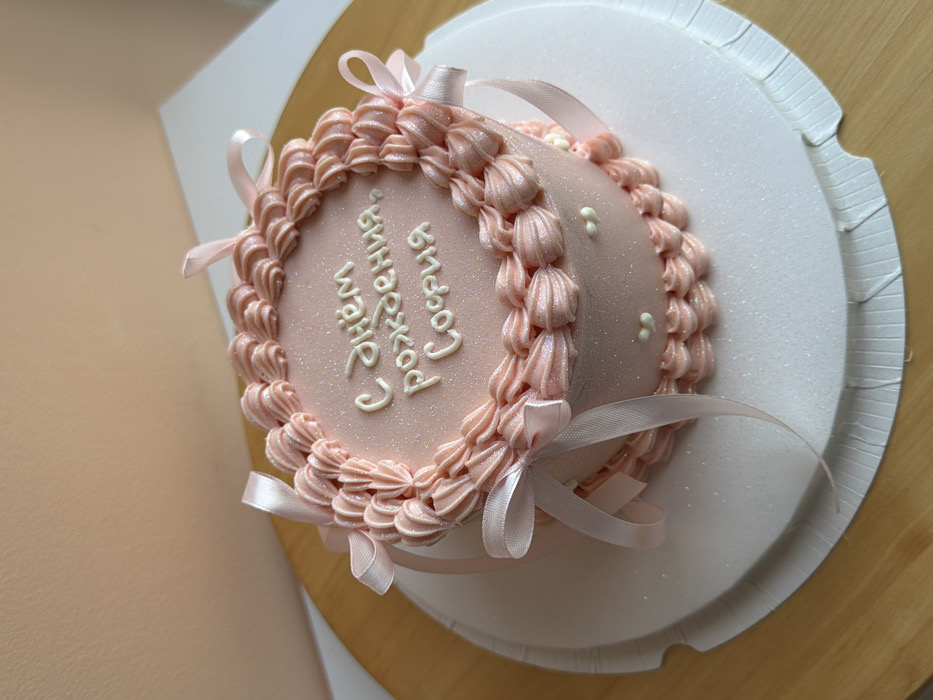 A round, pink cake is decorated with rosettes along the edge and white dots sparsely placed on the top. A pink ribbon is tied in bows around the cake’s perimeter. White cursive lettering is vertically oriented on the cake’s surface. The cake sits on a white, scalloped-edged cake board, which is on a wooden surface.