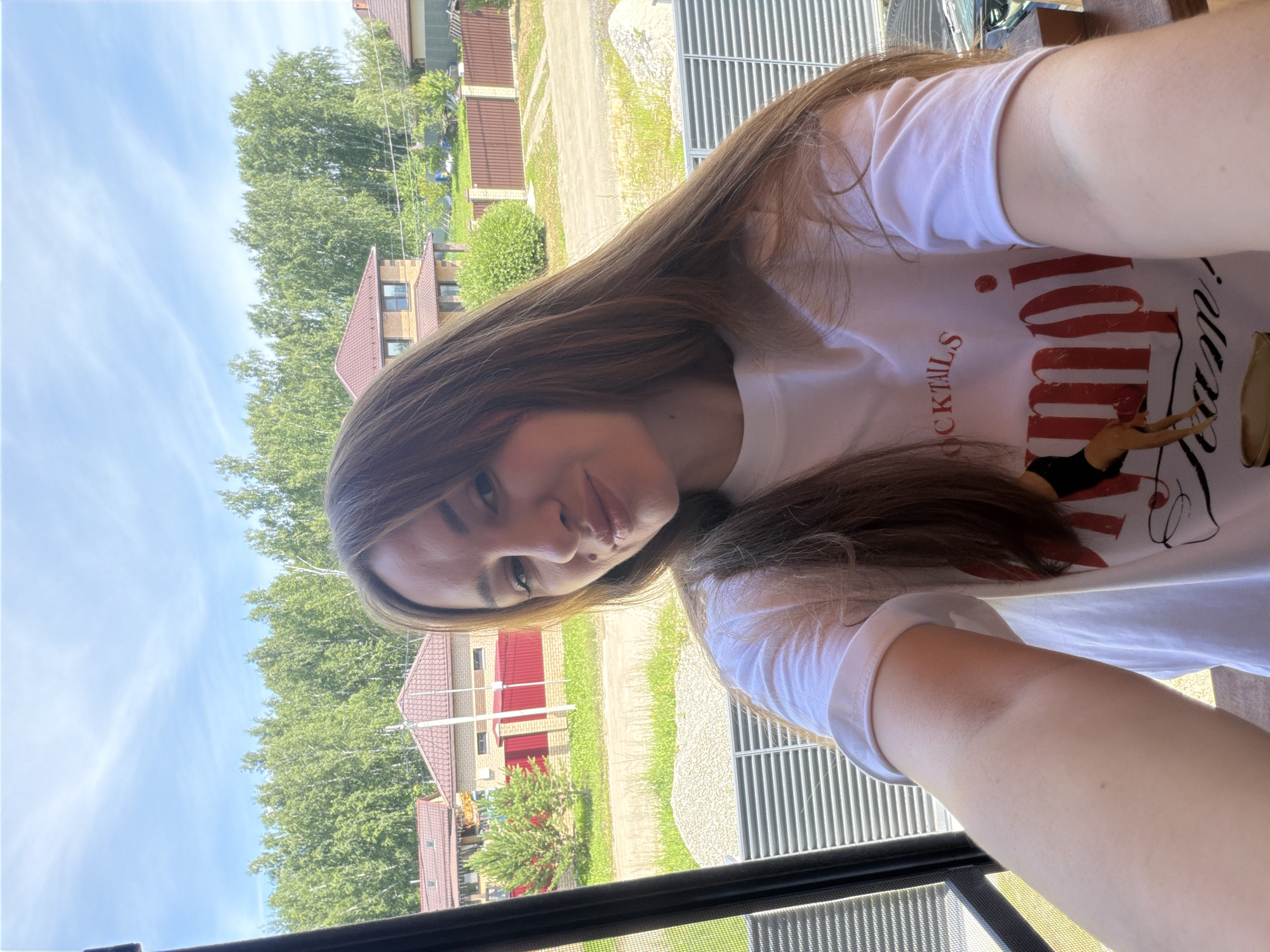 A young Caucasian woman with long, brown hair is taking a selfie outdoors. She is wearing a white t-shirt with red text and a graphic of a woman. The woman is in focus, and the background is blurred. Behind her, there are houses with red roofs, green trees, and a blue sky with white clouds. The image appears to have been taken on a sunny day. The location is in Malinovka, Russia.