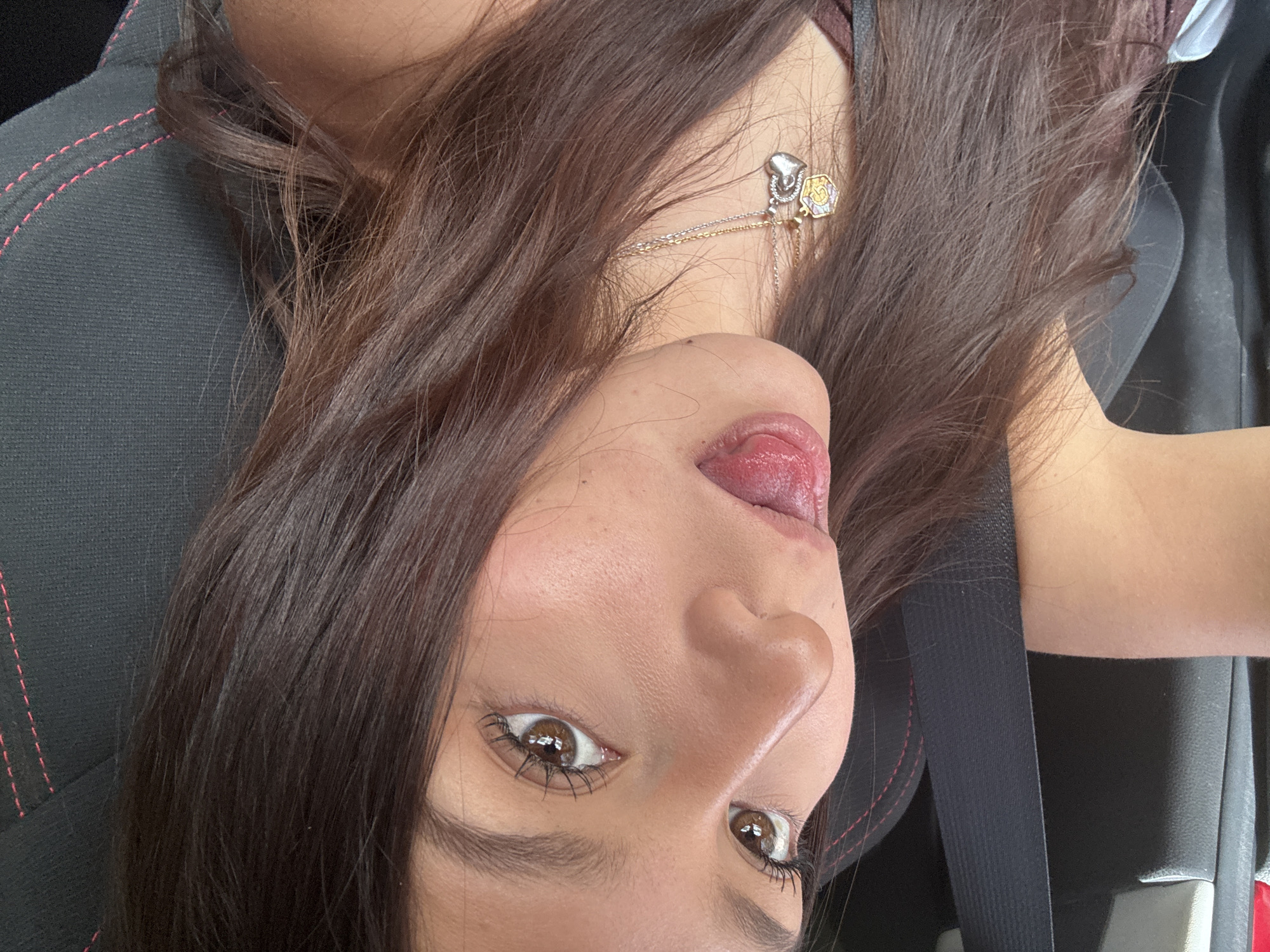 A woman with long brown hair is in the driver's seat of a car. She is sticking her tongue out. She is wearing a necklace. The car seat is black with red stitching. A seatbelt is visible. The location is Tlalixtac de Cabrera, Mexico.