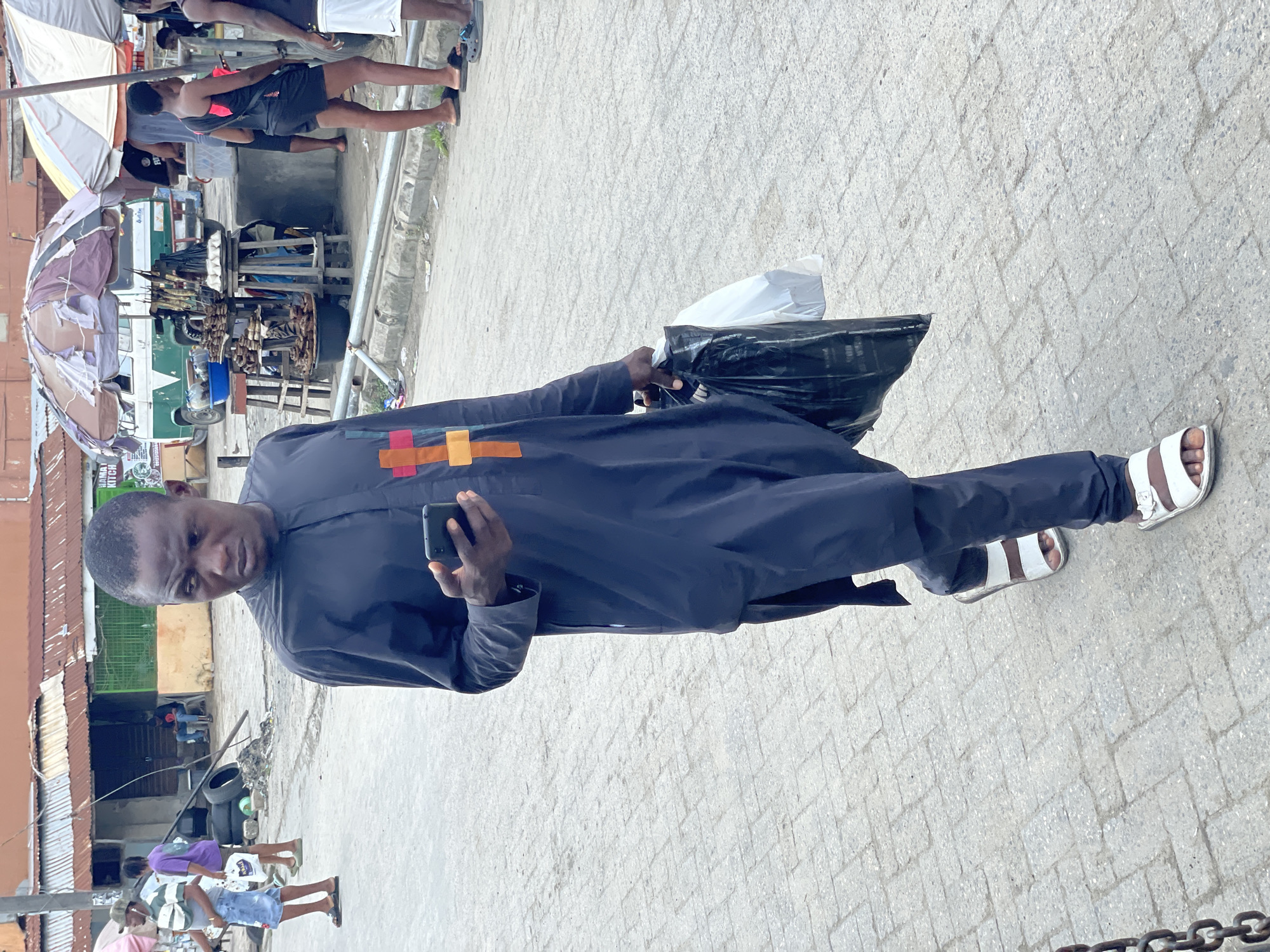 The image portrays a lively street scene, likely a market or busy thoroughfare, set in Ado, Nigeria. In the foreground, a man of dark complexion stands facing the viewer with a neutral expression. He is dressed in a dark blue or black long-sleeved tunic featuring a vertical band of colorful rectangular stripes (red, orange, yellow, green, light blue) on the chest. He wears dark trousers and white and brown open-toed sandals. In his left hand, he holds a black smartphone, and in his right hand, he carries a black plastic bag that encloses a white plastic bag.
The background reveals a bustling environment with several other people engaged in various activities. The ground is paved with light-colored interlocking bricks. To the left, there's a market area with makeshift stalls, some shaded by tattered umbrellas. One stall displays what appears to be goods on skewers, possibly smoked fish or meat, stacked alongside egg cartons. A white and green van is partially visible behind the market setup, and discarded tires lie nearby. Buildings with corrugated metal roofs and simple wall structures line the street. A sign, partially visible in the distance, bears the text "HANNAH KITCHEN." The scene suggests a typical daytime market atmosphere.