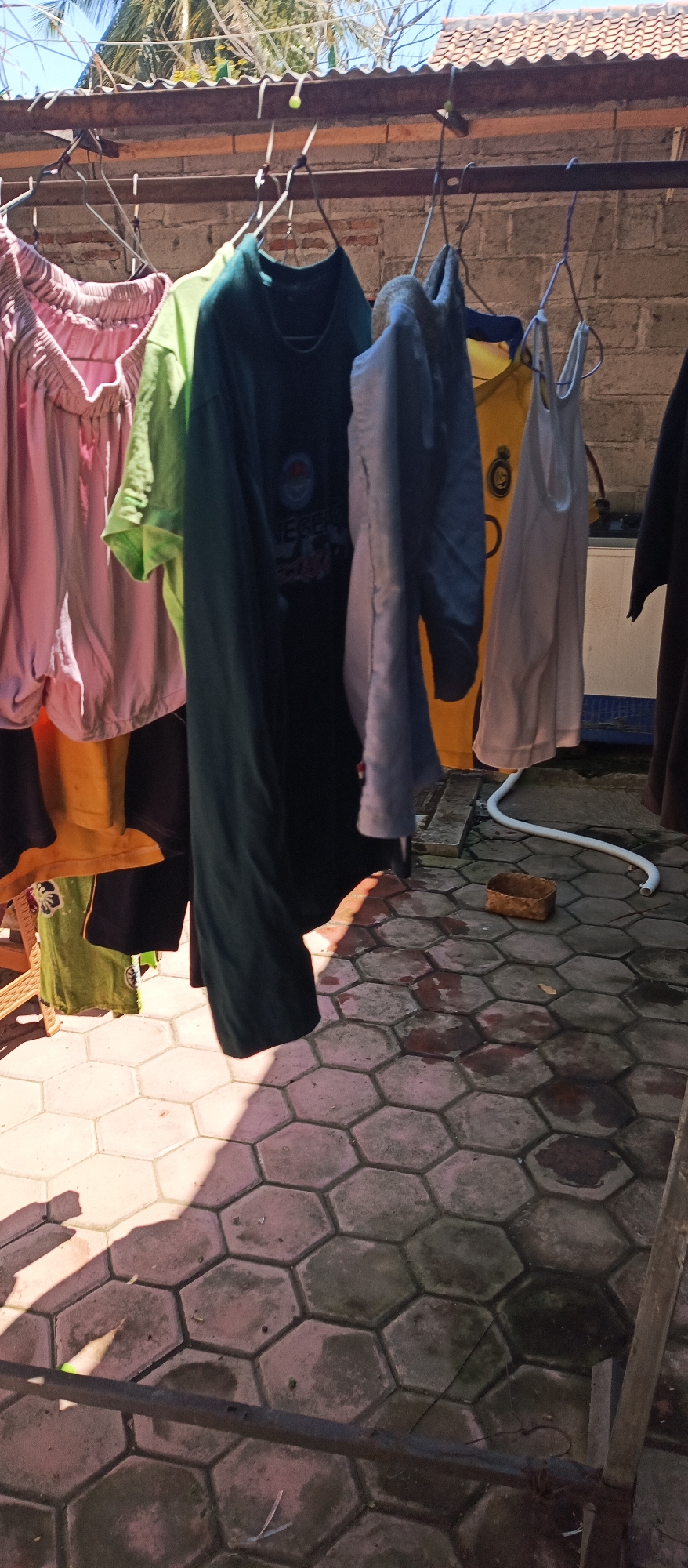 The image shows laundry hanging on a clothesline in what appears to be a backyard area. There are several items of clothing, including shirts and pants, suspended from metal hangers. The clothesline is strung between two horizontal metal poles, which are supported by a brick wall in the background. The ground is covered in hexagonal paving stones, and there is a white hose and a small woven basket on the ground. Sunlight casts shadows on the paving stones, indicating it is daytime. The overall scene depicts a typical domestic outdoor setting.