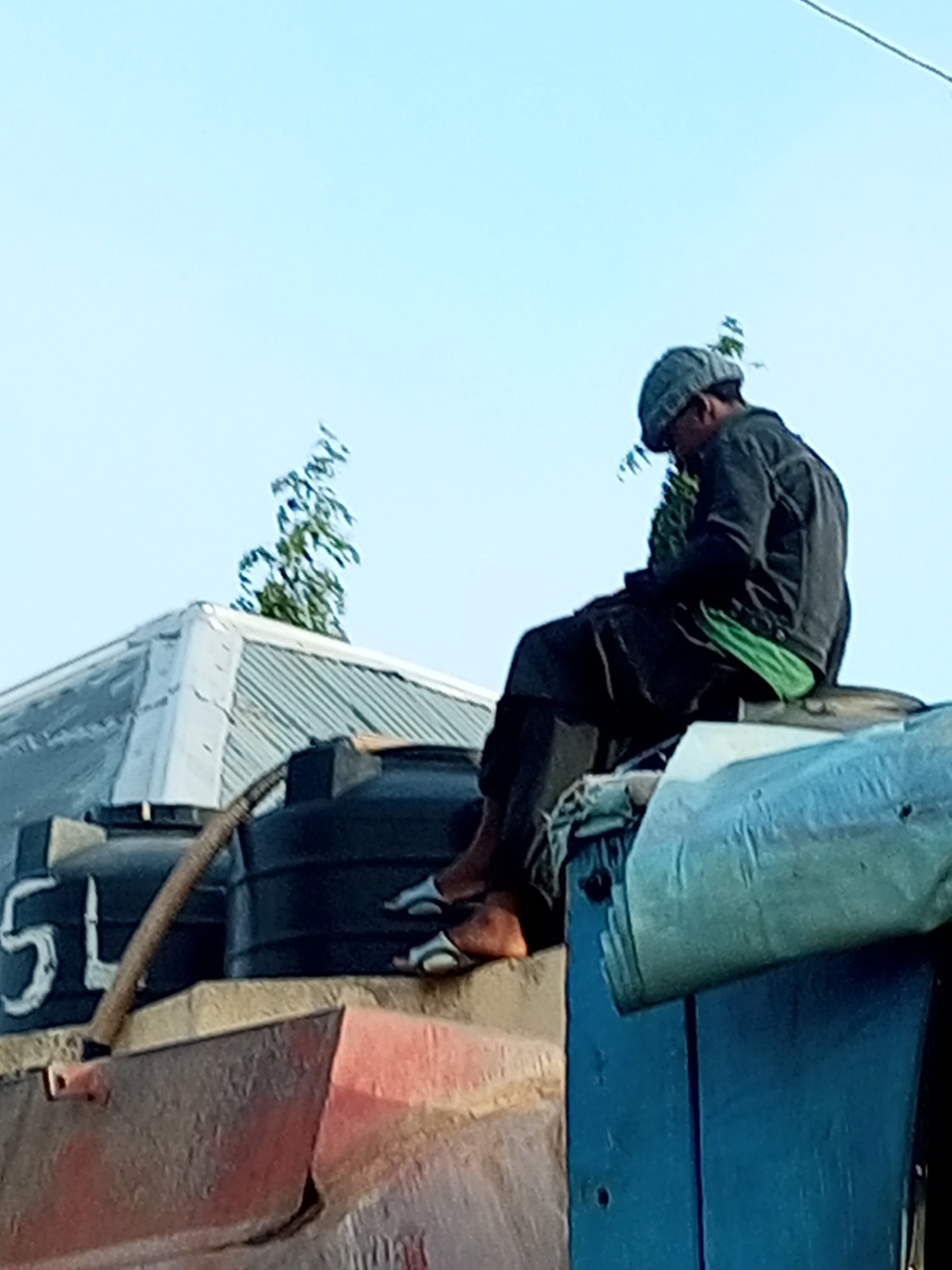 A person, possibly male, is sitting on top of a blue structure. The person is wearing a grey cap, a dark grey jacket, black pants, and green shorts underneath. They are also wearing white sandals. To the left of the person, there are two black water tanks with white markings. The front tank has the number "54" visible. A brown hose is attached to one of the tanks. Behind the water tanks, there is a corrugated metal roof of a building with a tree growing behind it. The sky is clear and blue. The scene appears to be outdoors, possibly in an informal settlement or industrial area. The location is specified as Tempure, Nigeria.