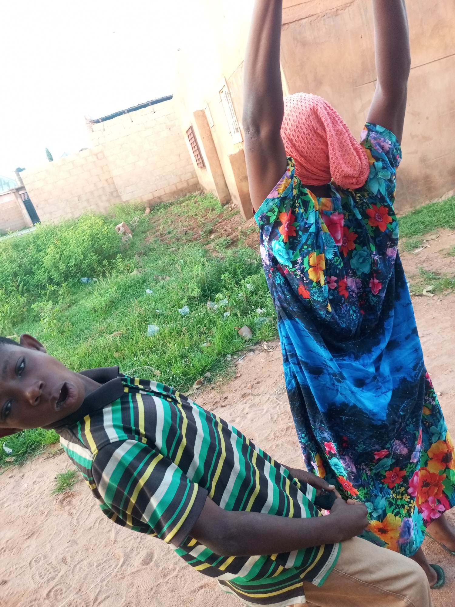 In Tempure, Nigeria, two individuals are captured in a slightly tilted, full-body outdoor shot. In the foreground, a young boy, likely of West African descent, is angled towards the camera. He is wearing a horizontally striped short-sleeved shirt in shades of green, black, yellow, and white, along with tan pants. His mouth is open in an expression of surprise or excitement. Behind him, a person, likely an adult, is facing away from the camera, their arms raised upwards. This individual is wearing a bright, floral-patterned blue dress with red, orange, yellow, and green flowers, and a pink headscarf. The environment appears to be a sandy, unpaved area with sparse vegetation and a brick wall in the background, suggesting a rural or semi-urban setting within Tempure. The overall lighting indicates it is daytime.