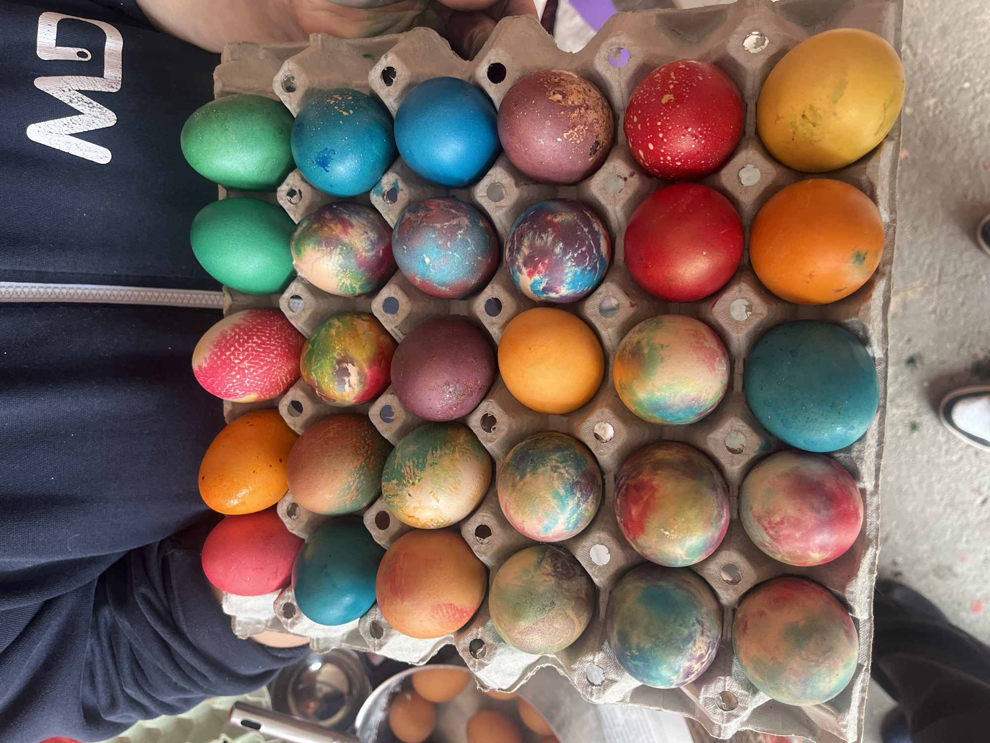 A person is holding a cardboard egg carton filled with approximately 30 dyed Easter eggs of various colors and patterns. The eggs are arranged in a 5x6 grid. The colors include green, blue, red, yellow, and orange, with some eggs exhibiting speckled or marbled designs. The person is wearing a dark-colored garment, possibly a jacket or sweatshirt, with white lettering. The background is slightly blurred, showing hints of other objects and surfaces, including what appears to be a kitchen counter, additional eggs in a bowl and a person's shoe. There is no information in the image connecting it to the city of Vlasatitsa, Bulgaria.