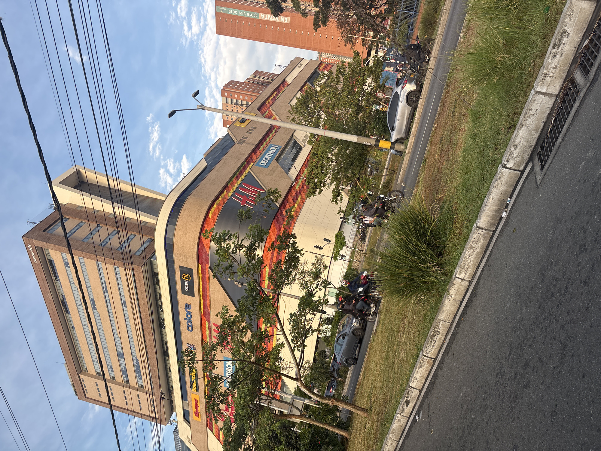 The image shows a street scene in Bello, Colombia. A multi-story building with several businesses including a Decathlon store is visible. Next to it is an office building identified as "Conconcreto". Cars and motorcycles are parked along the side of the road. There is a grassy area between the road and the building. Overhead power lines are present. The sky is blue with some clouds.