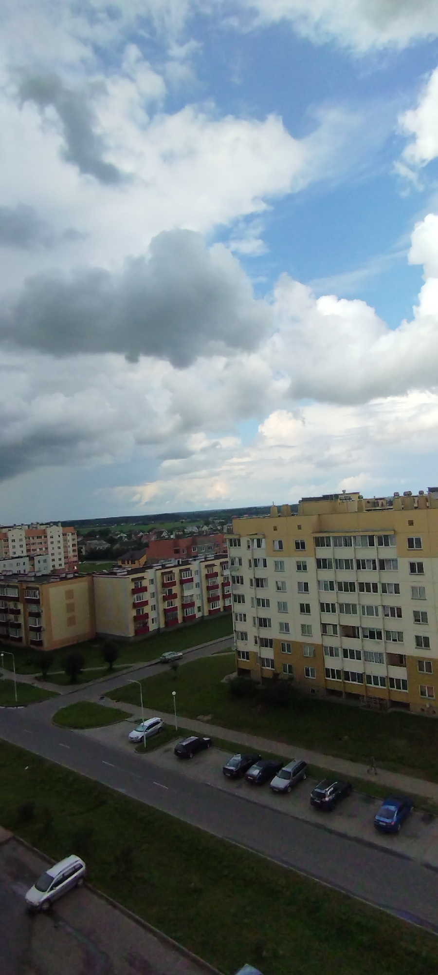 This is an aerial, medium-shot, outdoor photograph taken on a cloudy day in Skidzieĺ, Belarus. The image captures a residential area with multiple apartment buildings of varying heights and architectural styles. In the foreground, a paved road runs horizontally across the lower half of the frame, with several cars parked on the side and driving on it. A white SUV is parked on the left side of the road. A row of cars, including a dark sedan, a dark blue hatchback, and a silver sedan, are parked on the right side of the road, further down. Beyond the road, green grassy areas and sidewalks are visible.
Several multi-story apartment buildings dominate the middle ground. The building on the far right is a tall, yellow and white structure with numerous windows. To its left, a cream-colored building with red-framed windows and balconies is visible. Further left, more apartment buildings with similar red accents and cream or orange facades are arranged. In the background, a more distant cityscape of smaller buildings and houses stretches towards the horizon, bordered by a line of trees.
The sky above is filled with a dramatic display of clouds, with patches of blue sky peeking through. Darker, heavier clouds are visible on the left side of the frame, suggesting potential rain or a storm approaching, while lighter, fluffier clouds occupy the upper and right portions of the sky. The overall lighting suggests it is daytime, but the overcast conditions cast a diffused light over the scene.