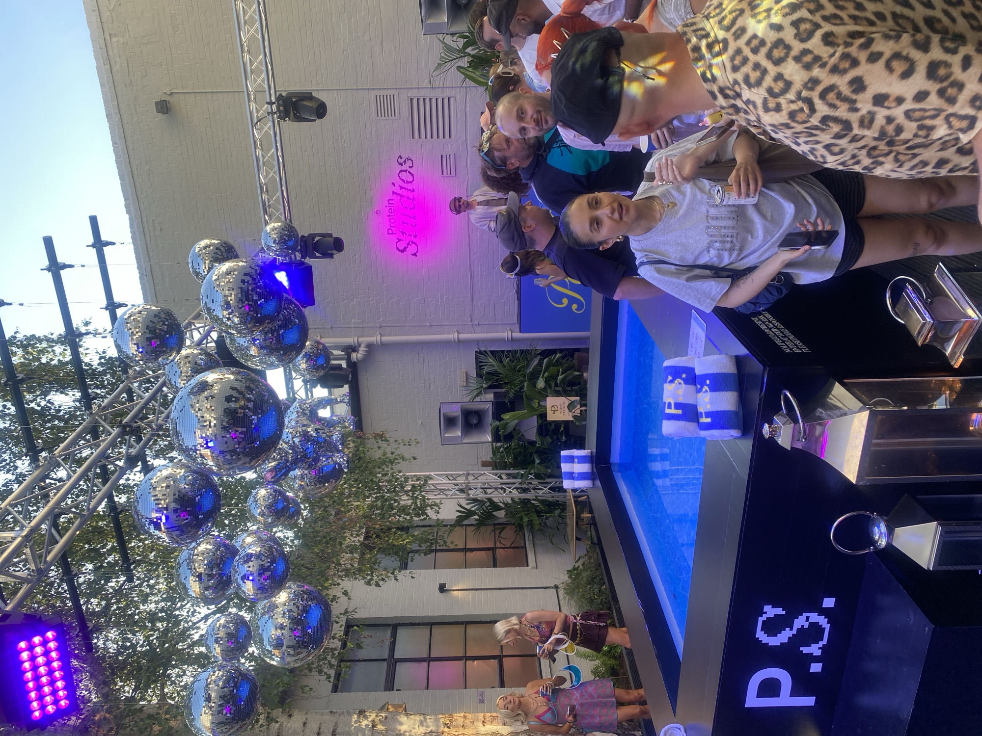 The image shows a crowded outdoor event in London, United Kingdom, likely a promotional gathering associated with "Protein Studios," as indicated by the neon sign. Several people are positioned near a small pool or water feature with branded towels. Disco balls hang overhead, adding to the celebratory atmosphere. Other branding elements include the "P.S." logo in pixelated lettering. Two women stand by the pool, seemingly engaged with something inside it. The environment suggests an urban outdoor venue.