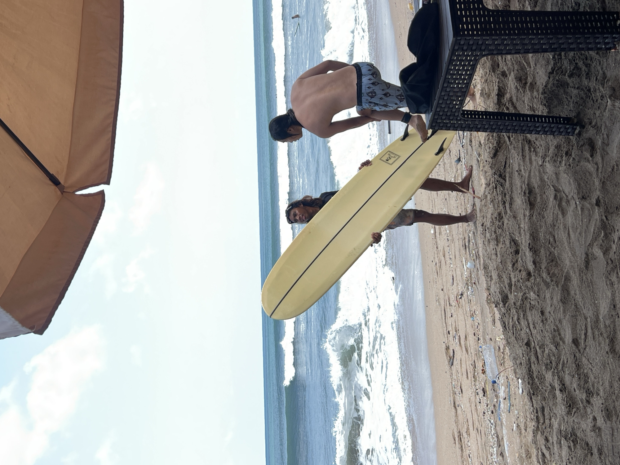The image depicts a scene on a sandy beach in Kuta, Indonesia, with the ocean in the background. Two men are standing near the shoreline. One man, on the left, is holding a pale yellow surfboard vertically. The other man, on the right, is bending over, with his back to the camera, appearing to adjust something on his leg or ankle, possibly a strap. He is wearing patterned swim shorts. The ocean has moderate waves with white foam. Part of a large, tan-colored umbrella is visible in the upper portion of the image, casting a shadow over the scene. In the foreground, a dark, woven plastic table is positioned on the sand. The beach appears to have some debris scattered on it. The sky is partly cloudy.