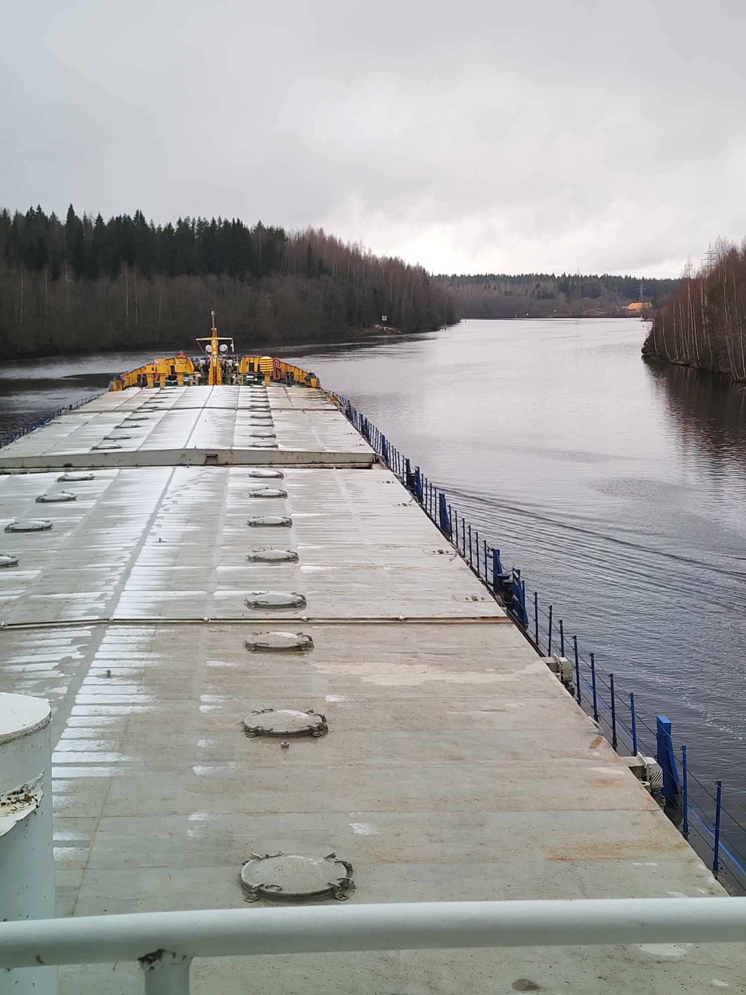 The image shows a view from the deck of a large barge or ship traveling through a narrow waterway. The deck is gray and covered in rectangular sections with circular hatches. A yellow structure, likely the bridge or command center of the vessel, is visible in the distance. Blue railings run along the side of the deck.
The waterway is surrounded by dense forest on both sides. The water is calm with slight ripples indicating movement. The sky is overcast with gray clouds.
The location is Depo, Russia.
