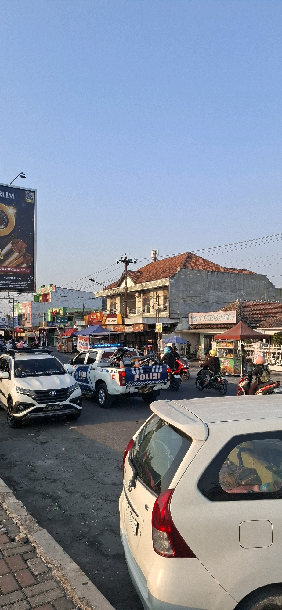The image depicts a street scene in Desa Ngesrep, Indonesia, likely during the late afternoon or early evening, as indicated by the warm, low-angled sunlight casting shadows. In the foreground, the rear of a white car is visible. Moving towards the middle ground, a white police truck with "POLISI" clearly marked on its side and tailgate is parked. The truck has blue flashing lights activated. On the truck bed, several motorcycles are being transported. A white SUV is parked to the left of the police truck.
Several individuals are present. A police officer appears to be standing near the rear of the police truck. Two individuals are on motorcycles in the traffic lane, one on a red motorcycle and another on a green motorcycle, both appearing to be moving. A person is also visible inside the white SUV. In the background, several buildings line the street, including commercial establishments with awnings and signage. One prominent sign reads "UMAYU KOSMETIK". Tents or stalls are set up on the sidewalk in front of some of the businesses, suggesting a market or food vendors. Power lines and poles are visible overhead. The sky is clear and blue. The road surface appears to be asphalt with some debris and oil stains. A concrete curb and sidewalk with paving stones are present along the side of the road.