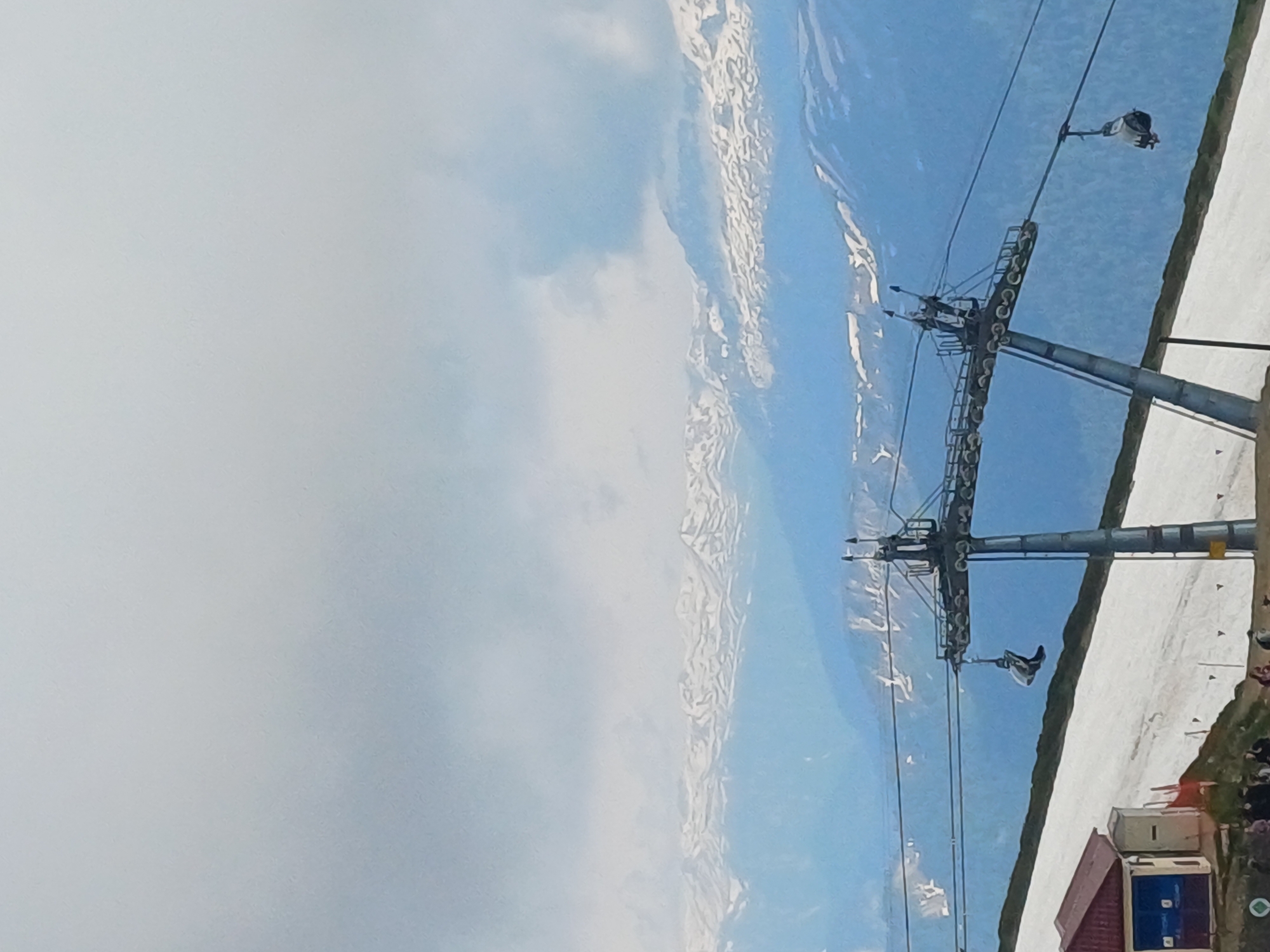 The image depicts a mountain landscape in Estosadok, Russia, dominated by a ski lift system against a backdrop of snow-capped mountains and a cloudy sky. A ski lift tower with attached cables and a gondola is prominently featured in the foreground. A single gondola is suspended from the cable, with its door open, appearing to be in motion. To the left of the tower, a small building with a red roof and blue doors is visible, situated on a snow-covered slope. Several people are gathered near the building, some standing and others appearing to be walking. The mountainous terrain extends into the background, with layers of snow-covered peaks fading into a hazy blue sky. The overall scene suggests a ski resort or mountain area in an operational state, possibly during the warmer months when snow remains on higher elevations.