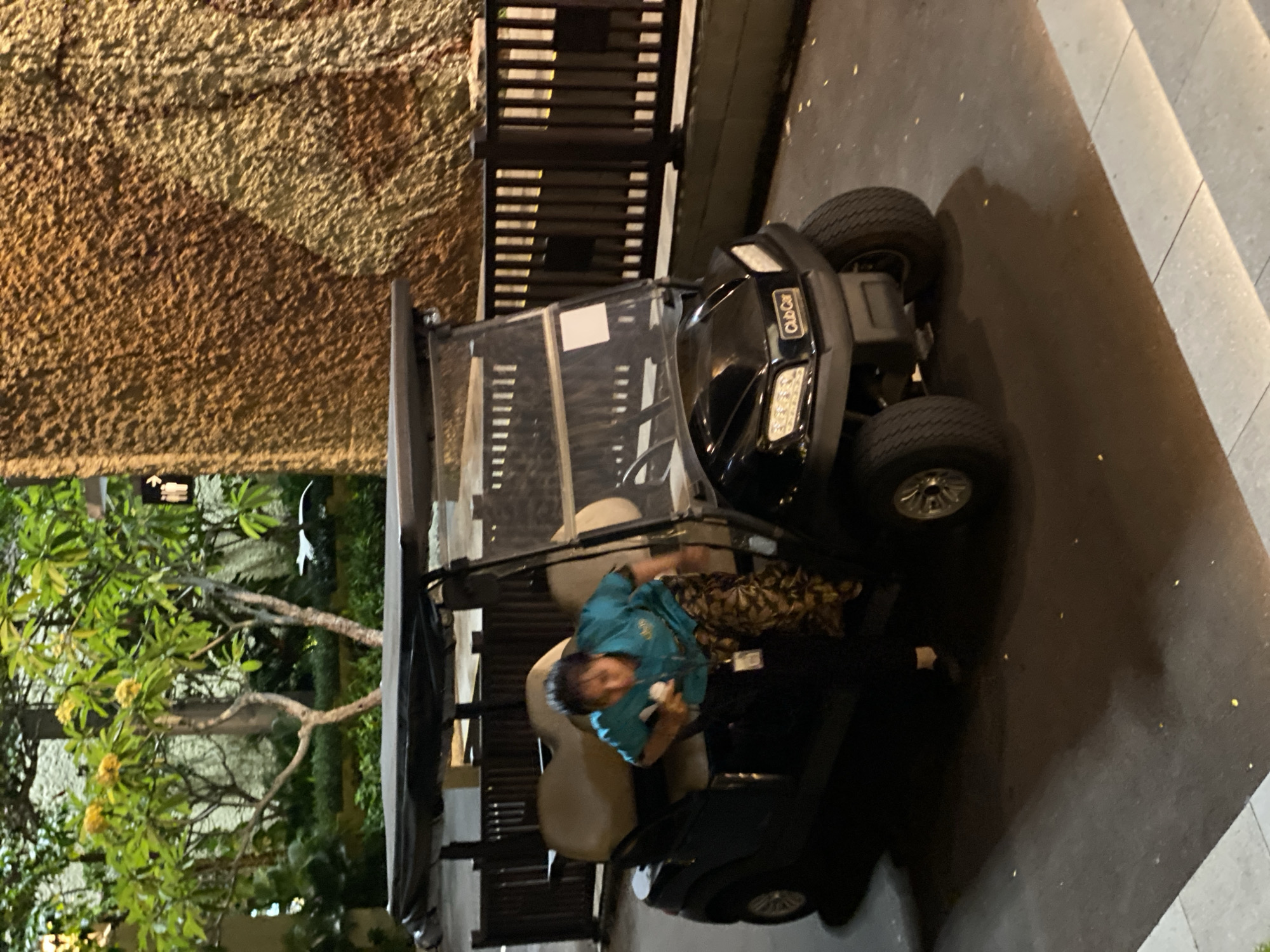 A person is getting into a black Club Car golf cart. The golf cart has a dark gray canopy and tan seats. The person is wearing a turquoise shirt and patterned pants, and is holding a white object. The golf cart is parked on a paved surface, with a dark brown wooden fence and a textured brown wall in the background. There are trees with green leaves and yellow flowers to the left of the golf cart. The scene appears to be outdoors at dusk or night, with artificial lighting illuminating the area. This location is consistent with Kuta Selatan, Indonesia, known for its resorts and golf courses.