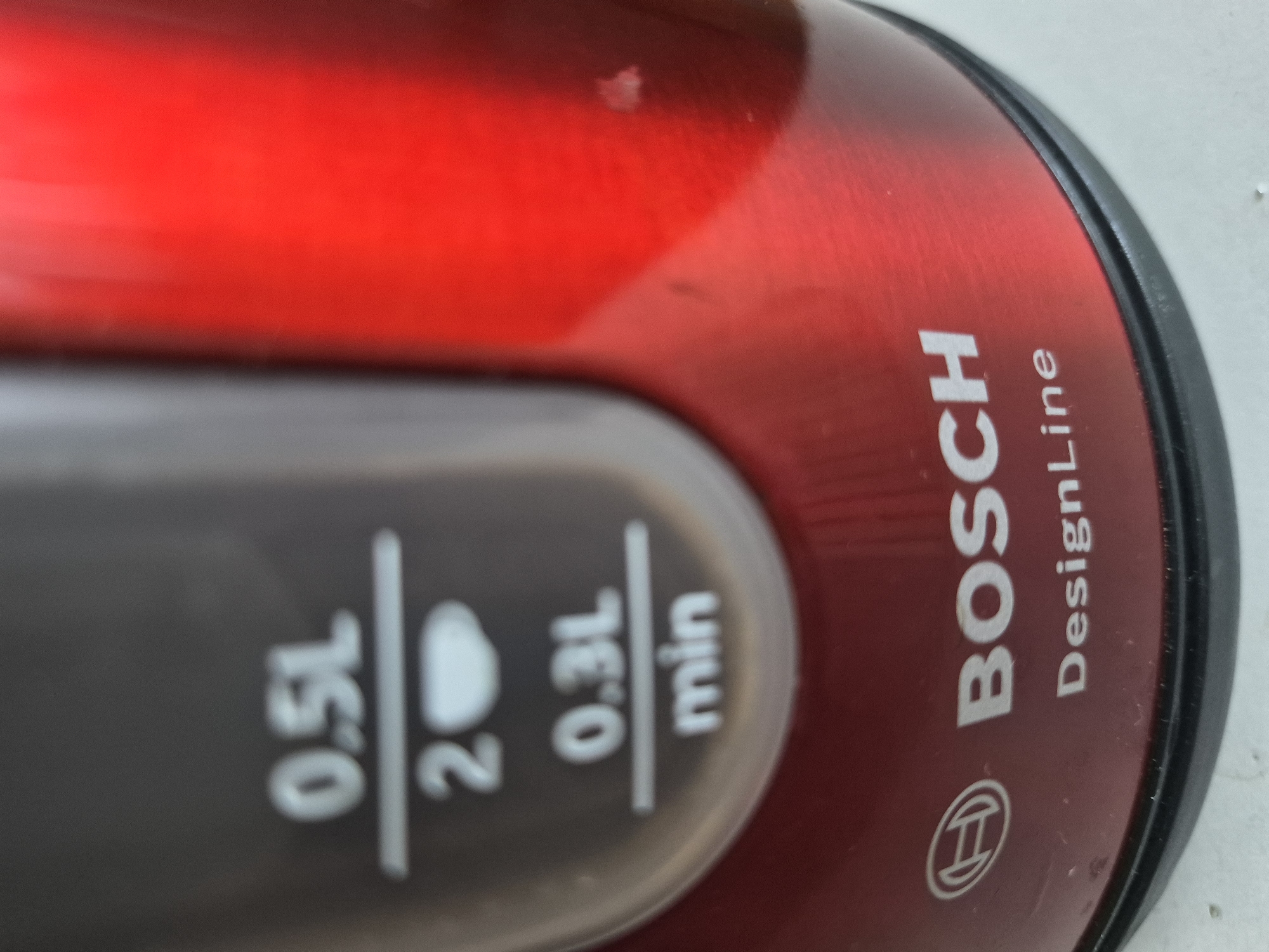 This is a close-up, angled shot of a red Bosch electric kettle. The kettle has a metallic, brushed finish with the "BOSCH" logo and "DesignLine" text printed vertically in white on the side. Below the logo, the Bosch symbol is visible.
On the side of the kettle, there is a water level indicator with markings for 0.5L, 0.3L, and a "min" indicator. There are also visual cues for 1.5L and 2, but the numbers themselves are not fully legible due to the angle and focus. The markings are white against a translucent grey background that is embedded in the red body of the kettle.
The overall lighting suggests an indoor setting, possibly a kitchen counter. There are no people visible. The image focuses on the branding and functional markings of the appliance.