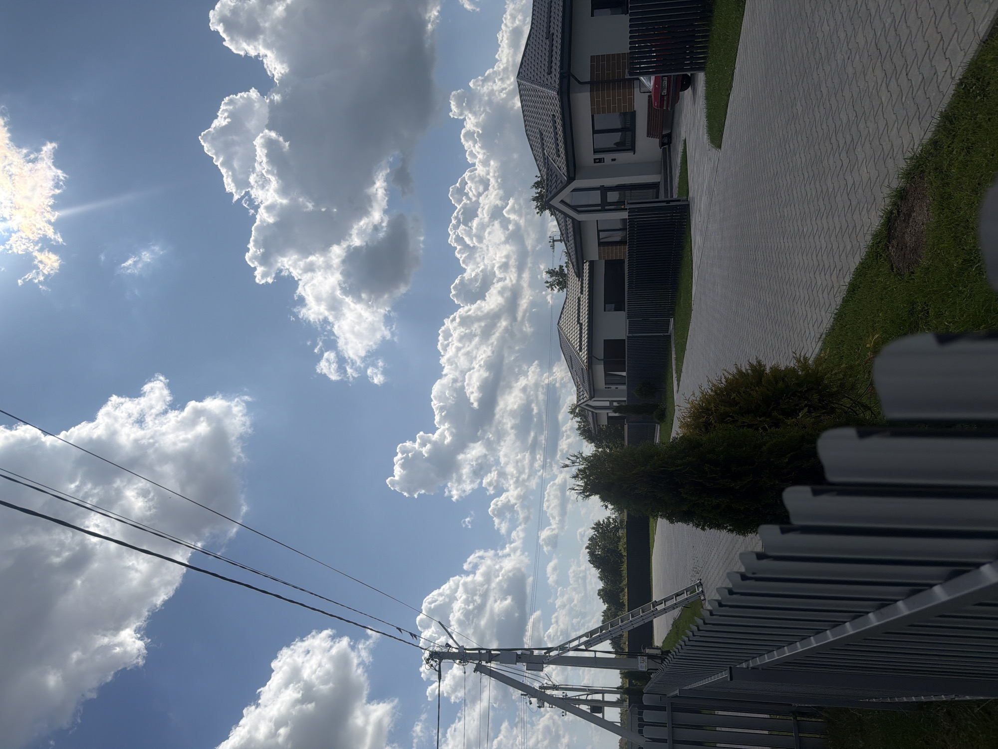 The image depicts a clear sky with fluffy white clouds and bright sunlight, suggesting daytime in a suburban or rural setting. In the foreground, a grey metal fence runs along the left side of the frame, bordering a paved driveway. Adjacent to the driveway are two modern houses with dark tiled roofs and light-colored walls, some featuring brick accents. One house has a prominent arched entrance with glass. A red car is parked in the driveway of one of the houses. To the left of the houses, a utility pole with wires and a ladder leaning against it is visible. Several evergreen trees are planted in the yards. The ground is covered with grey interlocking pavers, and there are patches of green grass. The scene appears to be a residential area, possibly in Golubevo, Russia, given the context.