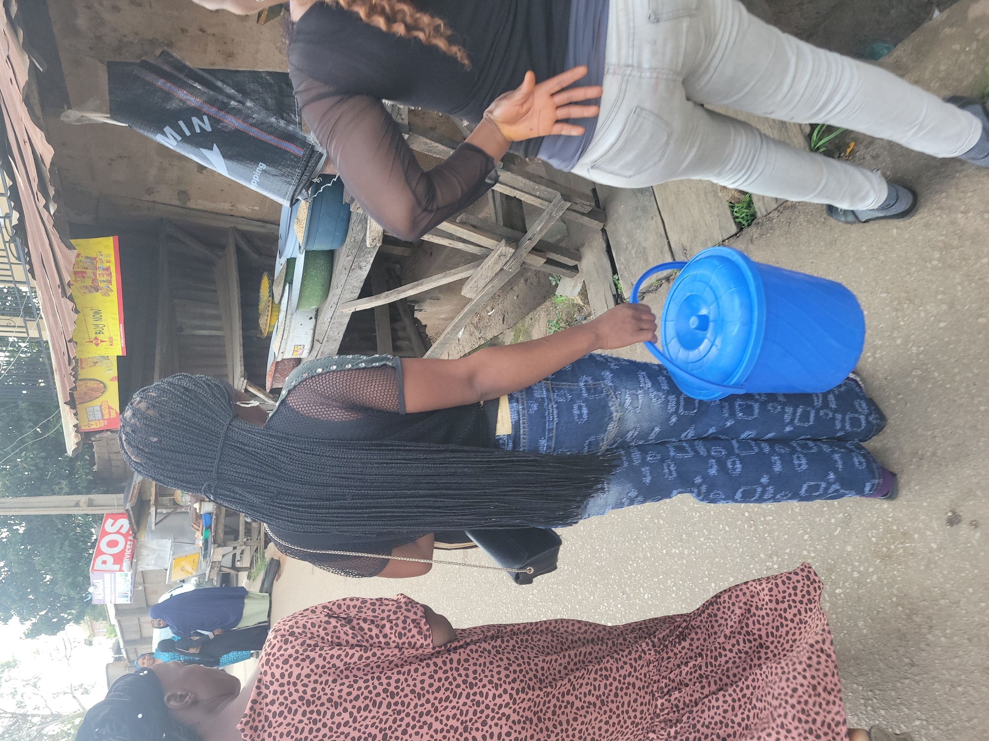 In a street scene in Jos, Nigeria, three women are observed. The woman on the left wears a brown and black spotted dress. The woman in the center has long, dark braided hair and is wearing blue jeans with a patterned design and a black mesh top. She carries a blue plastic bucket with a lid in her right hand, and a black purse with a gold chain strap is slung over her left shoulder. To her right, a third woman is seen from behind, wearing light-colored jeans and a sheer black top. In the background, a market stall is visible with wooden shelves. Signs on the stall include "POS SERVICES" and a yellow banner with red text that reads "BUY NOW MAGGI". Several containers holding what appears to be grain are arranged on a wooden table in front of the stall. The ground is paved with asphalt.