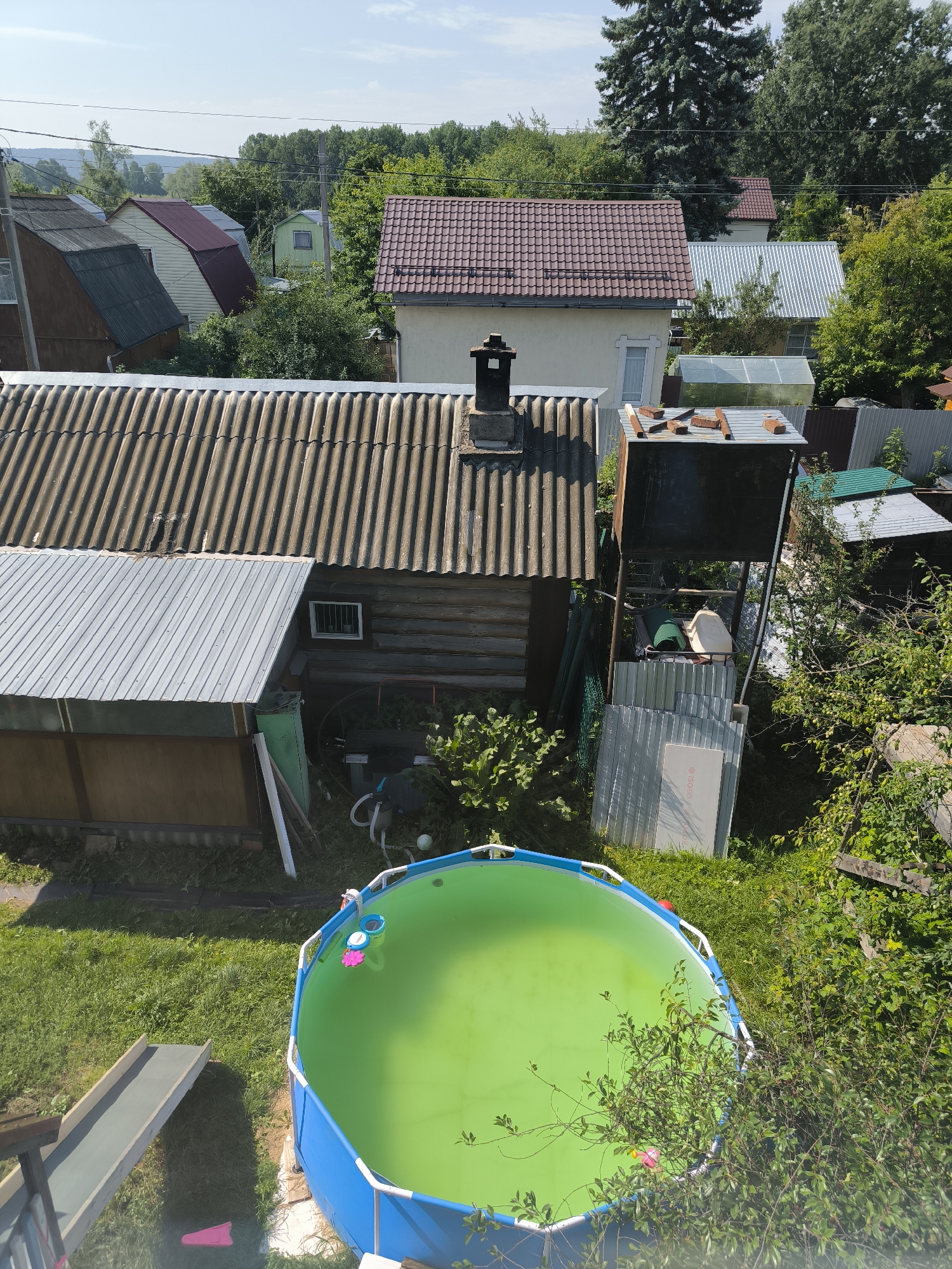 This is an aerial, high-angle, medium shot of a rural residential backyard in Oktyabrsky, Russia, on a sunny day. The scene is dominated by a bright green, circular, above-ground swimming pool with a blue frame, situated on a patch of lush green grass. A plastic slide, light gray in color, extends from the left edge of the frame towards the pool. In the background, several houses with various roofing materials, including corrugated metal and red tiles, are visible amidst dense green foliage and tall trees. A wooden structure, possibly a shed or outbuilding, is located to the left of the pool, with a corrugated metal roof. To the right of the pool, a metal platform structure with a dark colored tank on top and some stored materials underneath is present. The environment is characterized by a mix of residential buildings, overgrown vegetation, and a clear sky. No people or animals are visible in the frame.