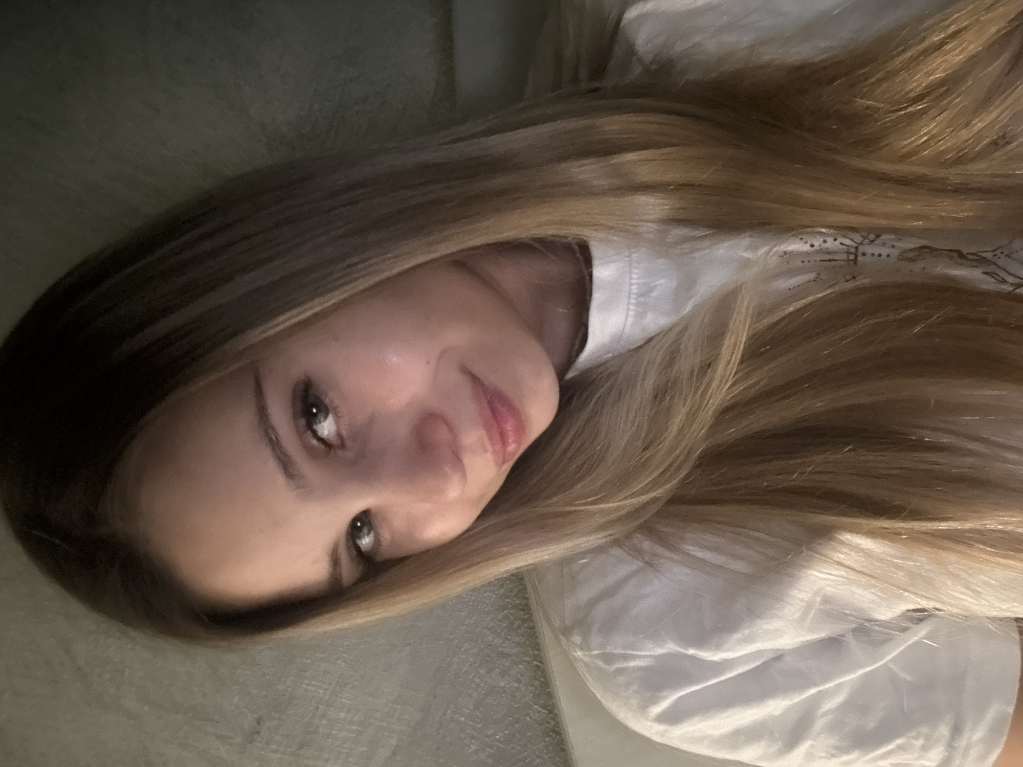 A close-up, head-and-shoulders selfie shows a young woman with long, blonde hair, parted down the middle. She is wearing a white t-shirt. Her head is tilted to her right, and she is looking directly at the camera. She has light blue eyes and a slight smile. The background is dark and textured, appearing to be a wall. The lighting is soft and angled, casting a shadow on the left side of her face. There is no indication of a specific location in Chaikovsky, Russia, within the image itself.