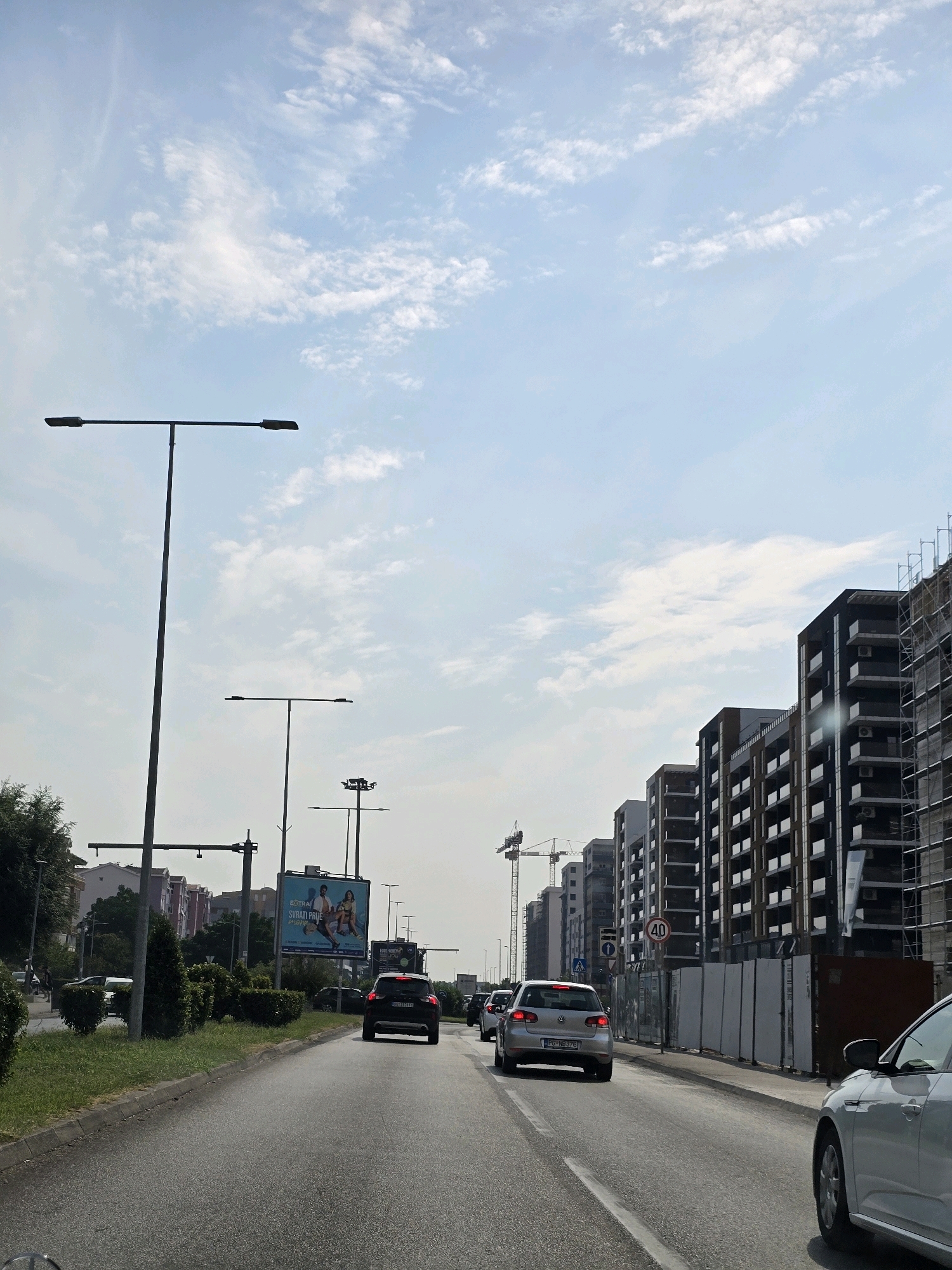 The image depicts a street scene in Podgorica, Montenegro, likely during daytime, with a clear sky and scattered clouds. The foreground shows a paved road with several cars traveling in the same direction. On the right side of the road, there are modern, multi-story buildings under construction, some with scaffolding. A speed limit sign indicating "40" is visible near the construction site. On the left side of the road, there are streetlights, a bus stop with a shelter, and a billboard advertising what appears to be a travel or leisure service. Further down the road, more residential buildings are visible, and trees and green areas line the roadside. The overall impression is of a developing urban area with ongoing construction and traffic.