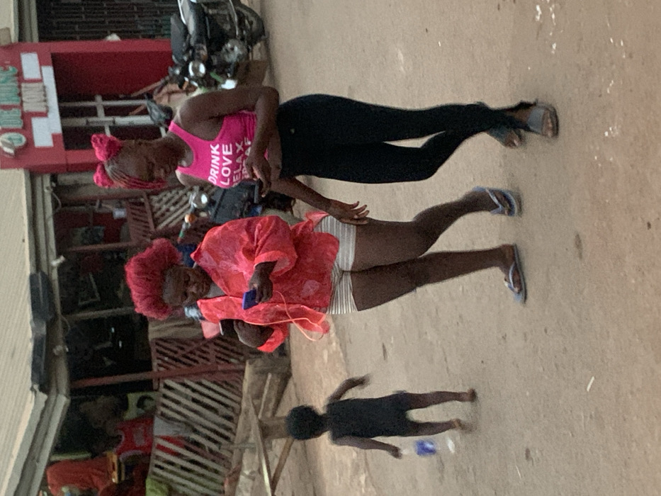 The image depicts an outdoor street scene in Otuocha, Nigeria, featuring three individuals: two adult women and a child. The lighting suggests it is daytime, possibly overcast.
In the foreground, a young child dressed in a dark outfit stands with their back to the camera. Behind them, a woman is seen wearing a bright red, flowing blouse over light-colored striped shorts, with light blue flip-flops on her feet. She holds a smartphone with white wired earbuds, seemingly engaged with the device. Her hair is styled in a distinctive red, curly or braided fashion.
Adjacent to her, another woman stands wearing a hot pink tank top, black leggings, and dark sandals. The pink top has white text printed on it, which clearly reads "DRINK LOVE RELAX". Her hair also appears to be styled or wrapped in pink. Her left hand is positioned near the arm of the woman in red.
In the background, a red building with a partial white sign (text illegible) is visible, along with what appears to be a parked motorbike or scooter. Other building structures with metal gates or grates are also present, indicating a commercial or residential area. The ground beneath the individuals is a light, dusty surface, likely dirt or concrete. The overall atmosphere is that of a casual, everyday moment in a local setting.