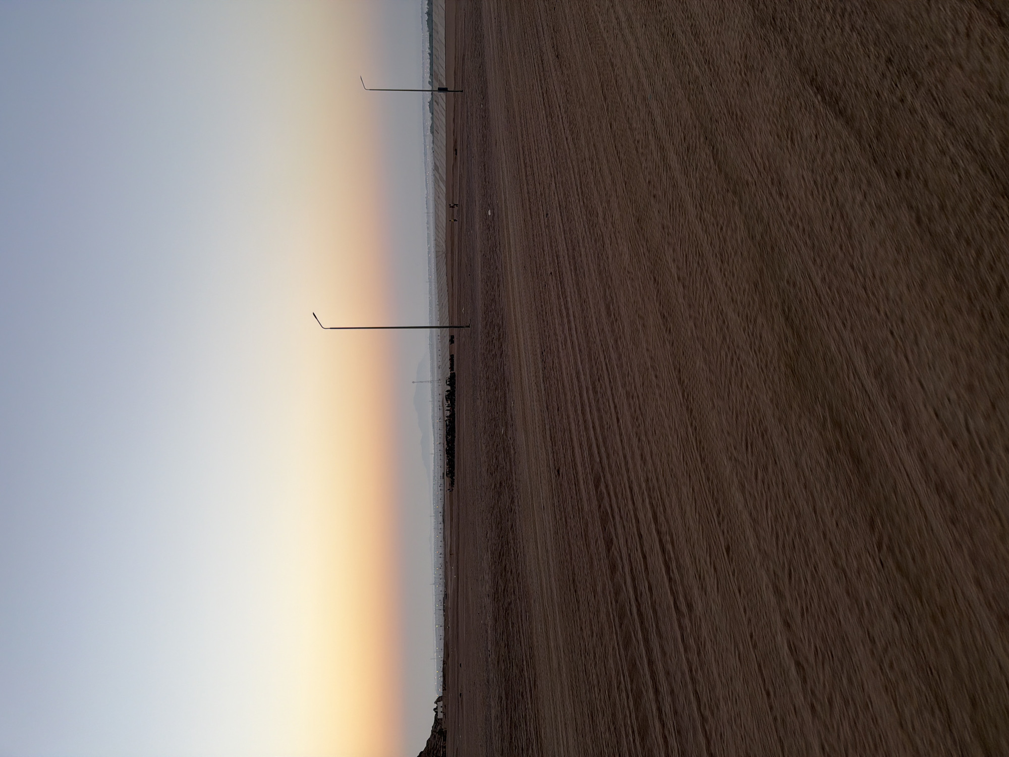 The image depicts a desert landscape at dusk, with a clear sky transitioning from pale blue at the top to a soft orange hue near the horizon. The foreground is a vast expanse of sandy terrain, marked with tire tracks. In the middle ground, a line of utilitarian streetlights stretches across the horizon, punctuated by several figures, some of whom appear to be walking. There are also a few dark, low-lying structures in the distance and a distant mountain range. The overall impression is one of a developing or sparsely populated area on the outskirts of a city. Specifically, the scene is identifiable as an undeveloped area of Sharm El Sheikh, Egypt, characterized by its arid environment and the characteristic desert illumination at the twilight hours.