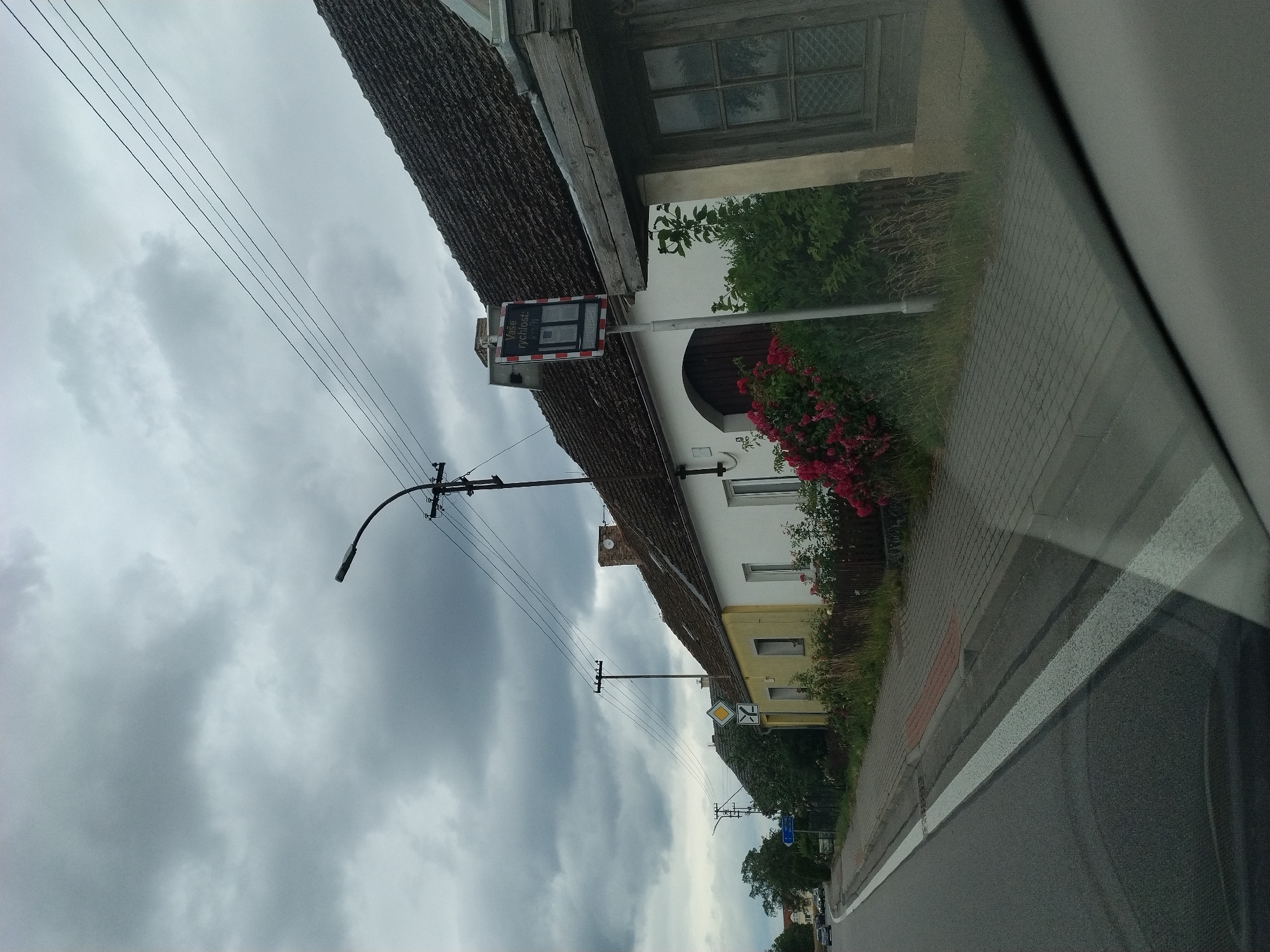 This image captures a street scene in Svatonice, Czechia, under an overcast sky. The photograph is taken from a vehicle, as indicated by the reflection on the right side of the frame.
The primary subjects are buildings and infrastructure. On the left, a traditional building with a dark, textured roof dominates the foreground. A street sign with text and a digital display is mounted on a pole near the building. The sign appears to be a speed display, with the text "Vaše rychlost:" (Your speed:) and a digital readout. Adjacent to this building, another structure with yellow and white facades is visible, with red flowers blooming in front of it. A sidewalk made of brick pavers runs alongside the buildings.
The scene is set on a road, with a grey asphalt surface and white lane markings visible. Several utility poles and power lines stretch across the frame, suggesting an inhabited area. Trees are also present, contributing to the somewhat rural or suburban feel of the location.
The weather appears to be cloudy and possibly damp, given the muted light and the appearance of the sky. The time of day is not clearly discernible, but the lighting suggests it is daytime. No people are visible in the frame, and the overall mood is neutral, focusing on the environment.
The visible text on the sign is "Vaše rychlost:".