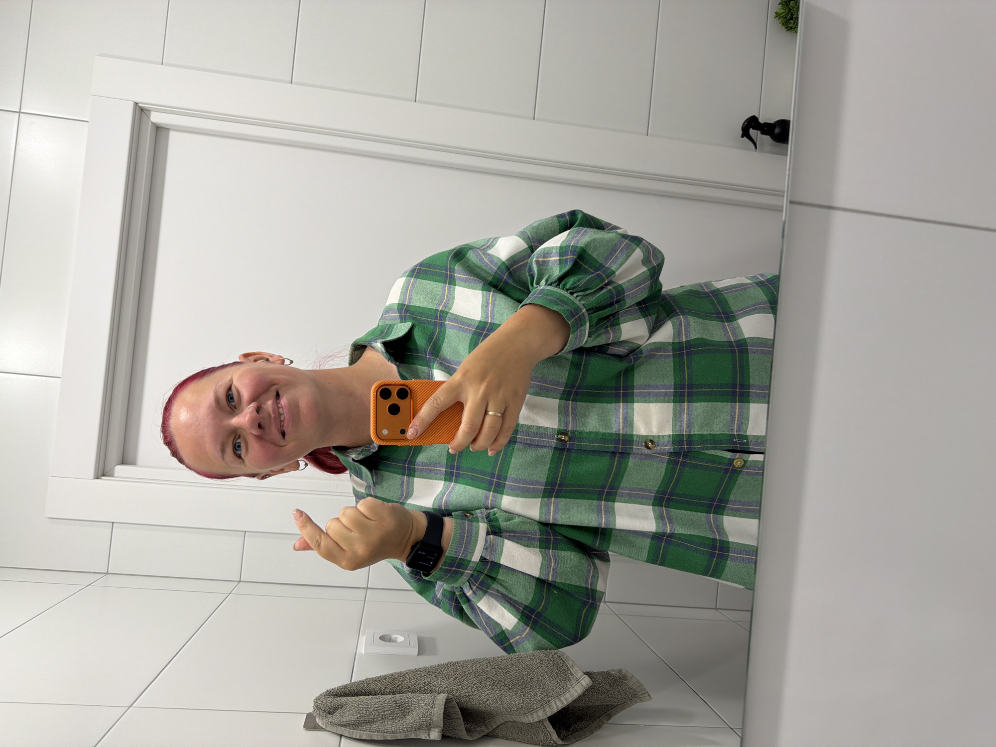 An adult female with red/pink hair, wearing a green, white, and blue plaid button-up shirt, is captured in a mirror selfie. She holds an orange-cased smartphone in her right hand, aiming it towards the mirror, and forms a Korean finger heart gesture with her left hand. A black smartwatch is visible on her left wrist, and a gold-colored ring is on her left ring finger. She has blue eyes, a nose ring on her left nostril, and hoop earrings in both ears, smiling directly into the camera.
The reflected background reveals a room predominantly featuring white square wall tiles with white grout. To the left, a grey towel hangs from a silver hook, and a white electrical outlet is mounted below it. To the right, a white door with a simple white frame is visible. Directly below the mirror's edge, a white shelf or counter displays a black spray bottle and a small, green artificial plant.
Based solely on the visual content of the image, there are no discernible features, landmarks, or specific indicators that allow for forensic identification of the location as Chaikovsky, Russia.