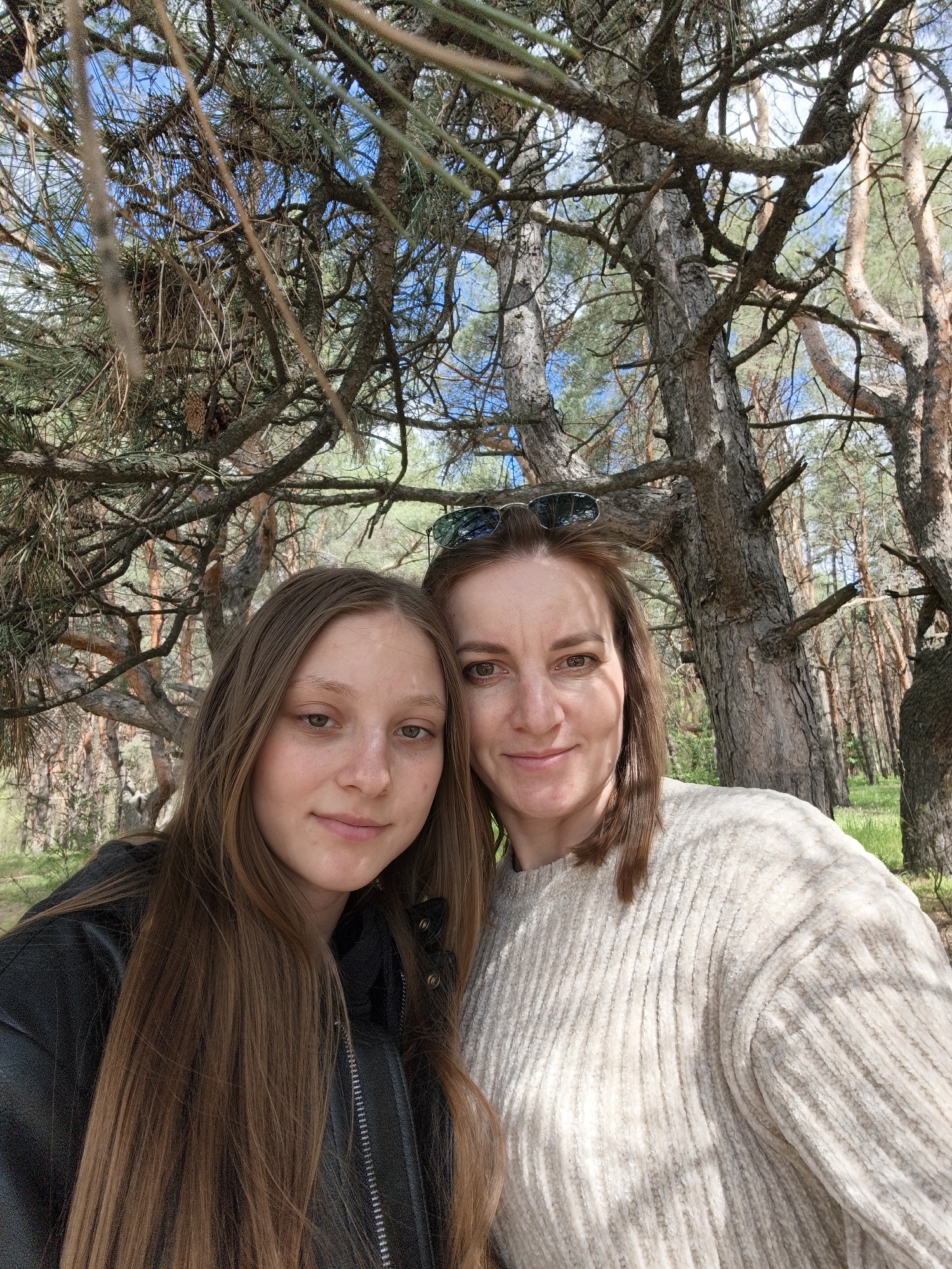 The image is a close-up, selfie-style photograph of two women in a pine forest, taken during bright daylight under a clear blue sky.
The subject on the left is a younger woman with long, straight brown hair, wearing a black leather-like jacket. She looks directly at the camera with a soft, pleasant expression. Beside her on the right is an older woman with brown, shoulder-length hair and sunglasses resting on top of her head. She also smiles faintly at the camera, wearing a cream-colored, ribbed knit sweater.
The background is dominated by the tall, textured trunks and branches of numerous pine trees. Pine needles and scattered pinecones are visible throughout the dense canopy, through which patches of blue sky are discernible. Sunlight filters through the branches, creating dappled shadows, particularly noticeable on the older woman's sweater. The overall setting suggests an outdoor activity or leisurely moment in a natural environment.
The location is Shyroke, Ukraine. There is no visible text in the image. Both individuals appear content.