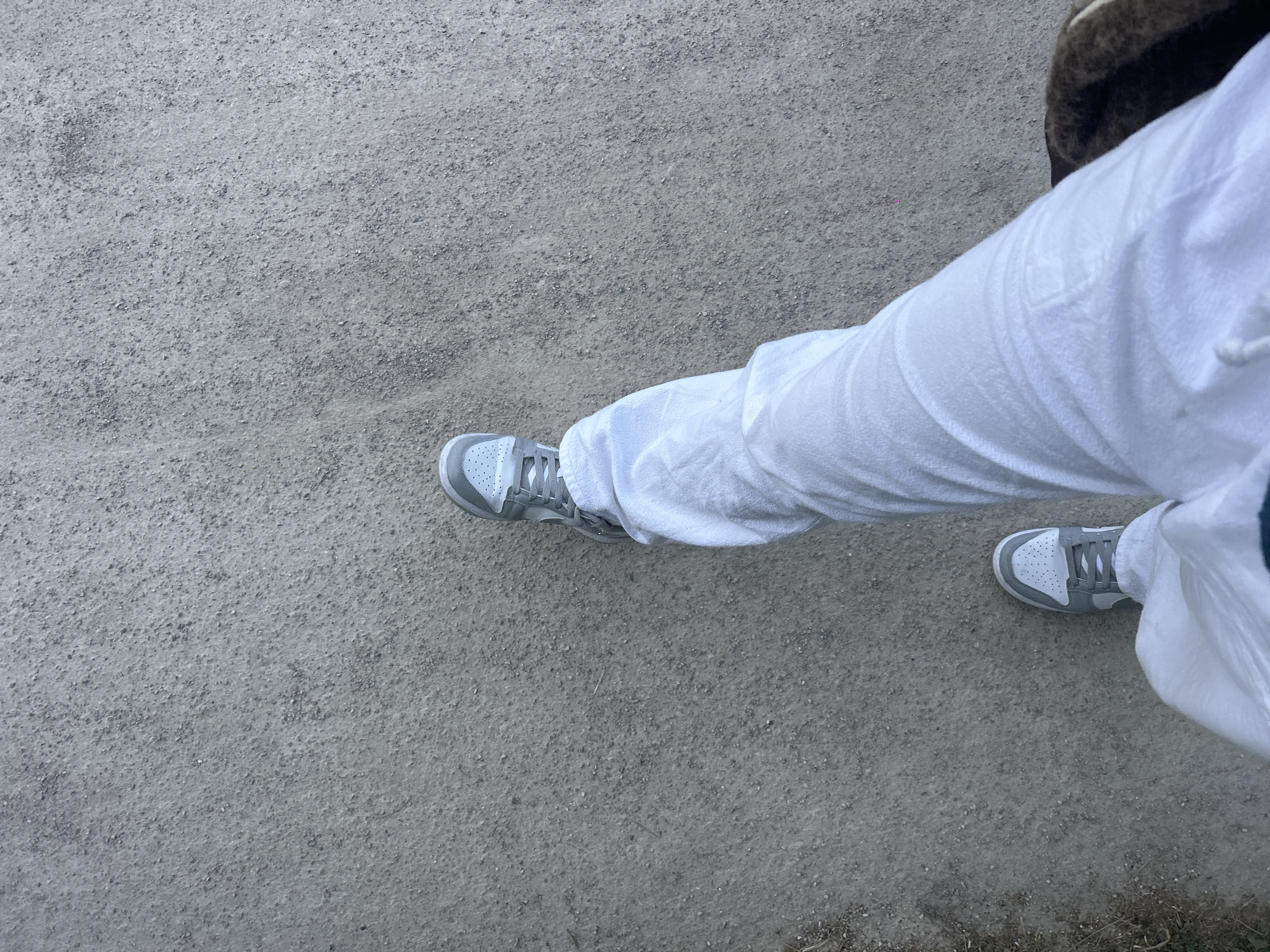 A downward-angled shot depicts a person's lower body, specifically the legs and feet, on a gray gravel surface. The individual is wearing light gray sneakers and loose-fitting white pants. The setting appears to be an outdoor area with a gravel ground. The environment suggests an urban setting, such as a park path or sidewalk. There is no information present within the image to suggest that the location is specifically in Marquette-lez-Lille, France.