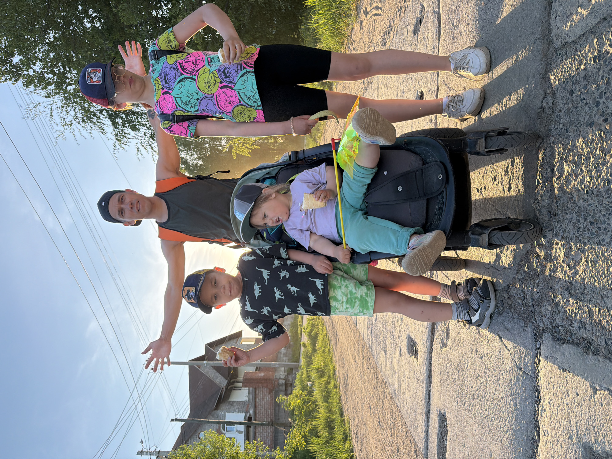 Four people are standing on a paved surface with a stroller. One child is seated in the stroller eating. The other people are positioned around the stroller. Two of them, a child and an adult, are making gestures with their hands. All four are eating or have eaten something from a cone-shaped pastry in their hands. There are buildings, vegetation, and utility poles in the background. The location may be in the city of Chaikovsky, Russia.