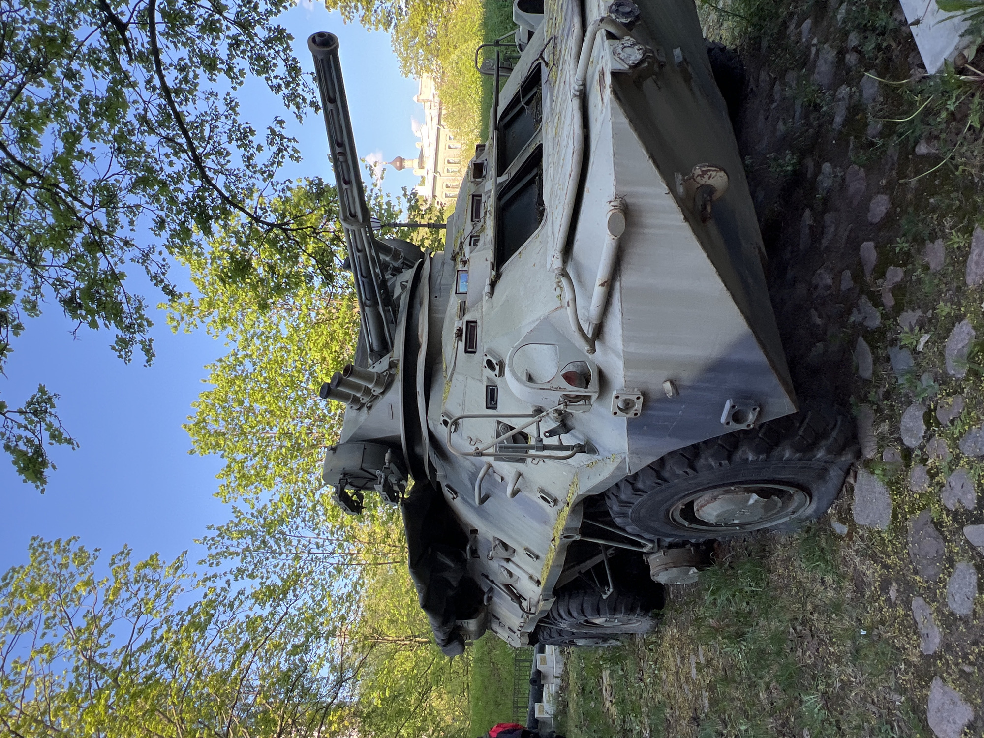 Here is a description of the image:
The image shows a BTR-80 armored personnel carrier on display outdoors. The vehicle is painted in a camouflage pattern and shows signs of wear. It is surrounded by grass and trees. In the background, a building with a gold dome is visible, indicating that the location is likely a park or public space in Saint Petersburg, Russia.