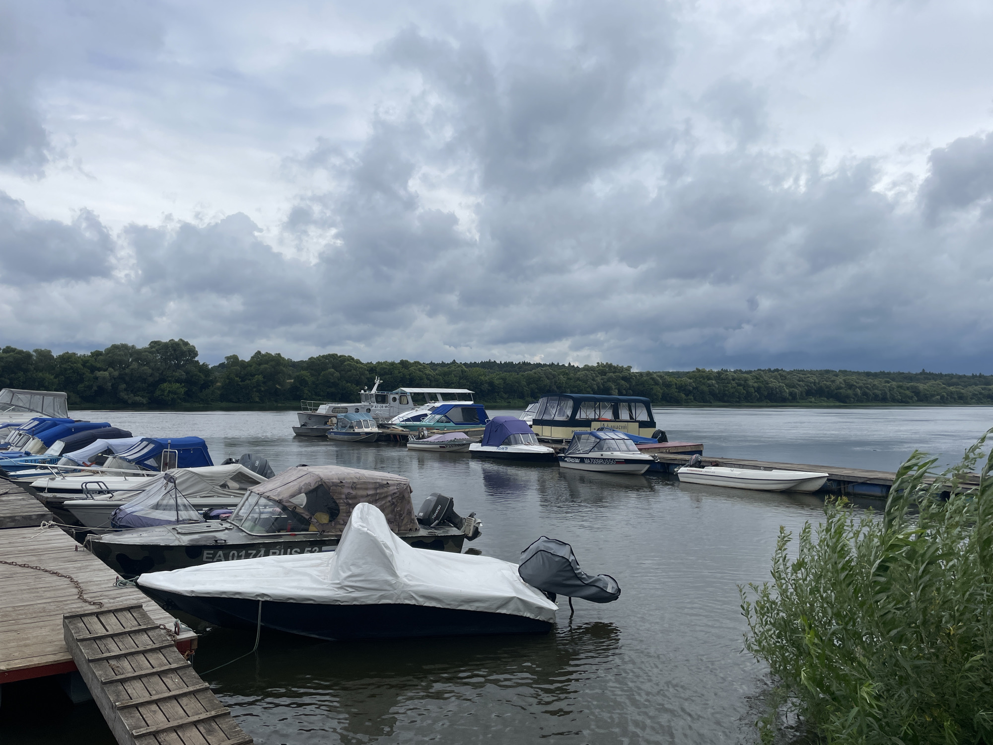 The image shows a cloudy day at a marina on the Oka River in Pushchino, Russia. Several boats are docked, some covered and some uncovered. A wooden pier is visible in the foreground, with a small ramp leading down to the water. The river is calm, and the opposite bank is lined with trees. The sky is overcast with heavy, grey clouds.