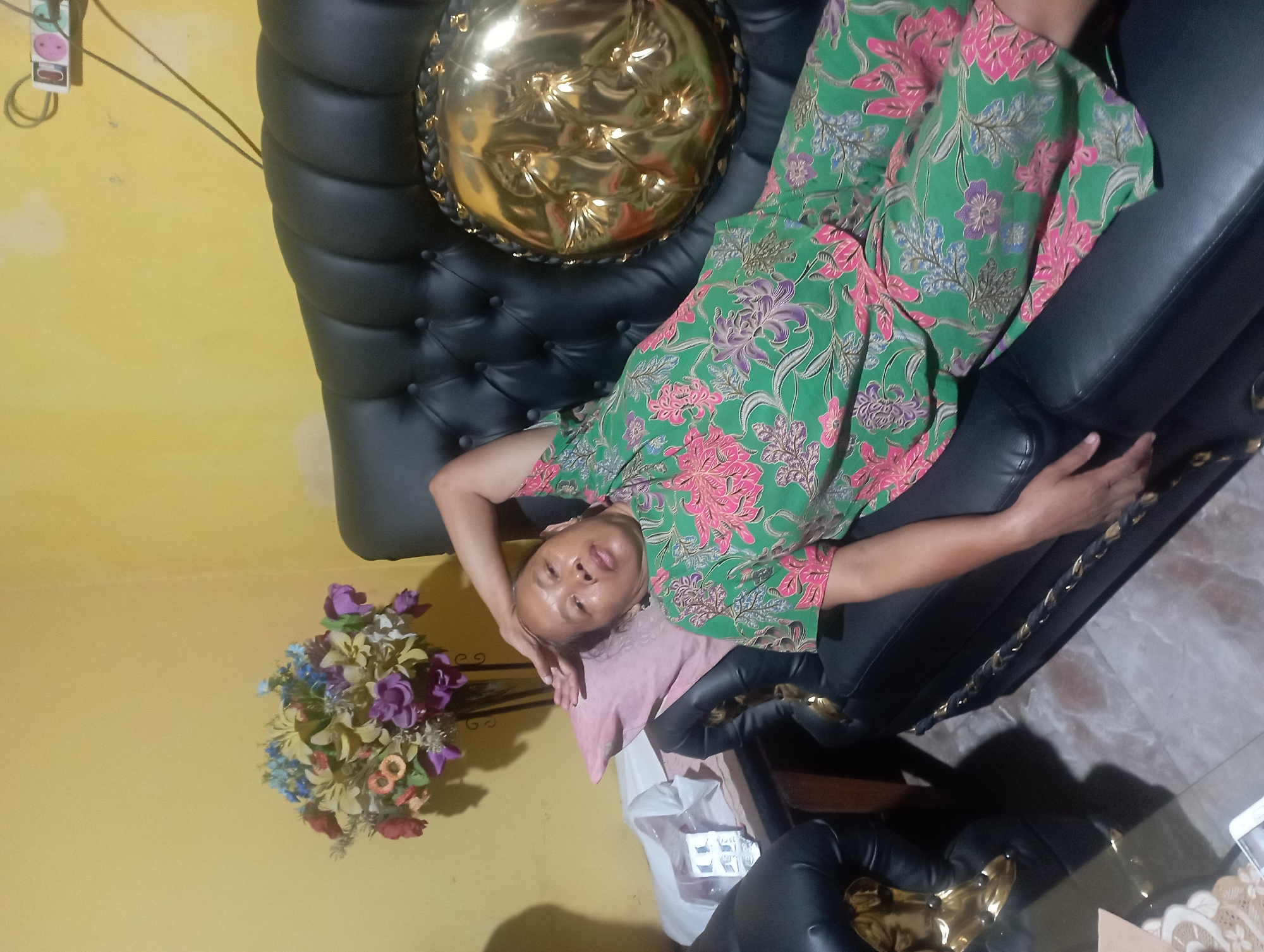 A woman is lying on her back on a black leather couch, her head resting on a pink pillow. She is wearing a green and pink floral-patterned outfit. Her right arm is bent and her hand is resting on her forehead. The couch is tufted and has a circular gold-colored decorative item on the right side. Behind the couch, the wall is painted yellow. To the left of the couch is a small table with a vase of artificial flowers and some items. A power strip with three outlets is visible on the wall above and to the right of the couch. The floor appears to be tiled. The overall impression is of a domestic indoor setting, potentially in Maos Kidul, Indonesia, given the context.