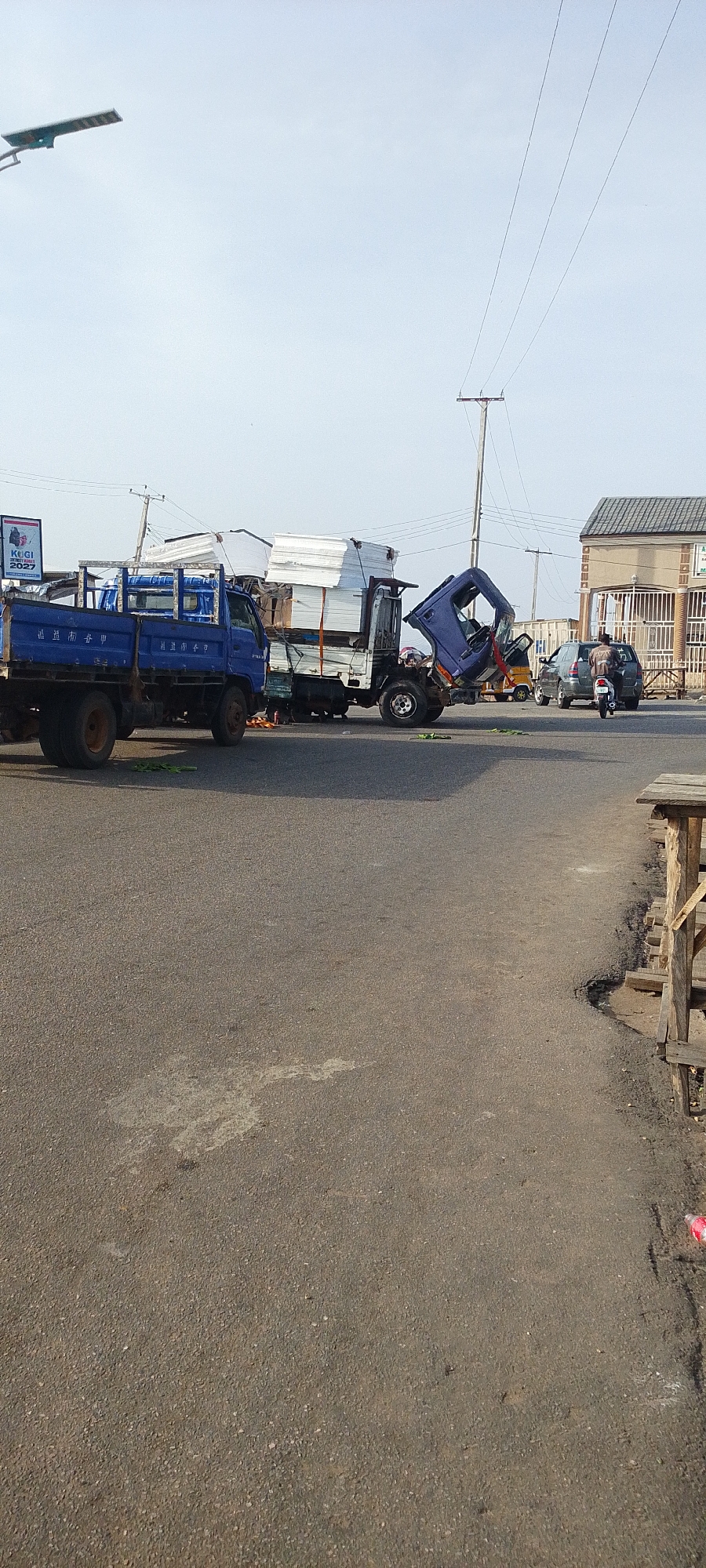The image shows a road traffic collision involving three trucks in Lokoja, Nigeria. One blue truck is loaded with green leafy material. A second truck is carrying a large, white, rectangular cargo. A third, blue truck appears to have its cab detached and tilted upwards, resting on the cargo of the second truck. Debris is scattered on the road. There are a few other vehicles including a motorcycle and a van or SUV. People can be seen standing near the accident scene. Overhead powerlines are visible. A building is in the background with fenced windows. A political poster is attached to a pole in the background. The location appears to be a road junction.