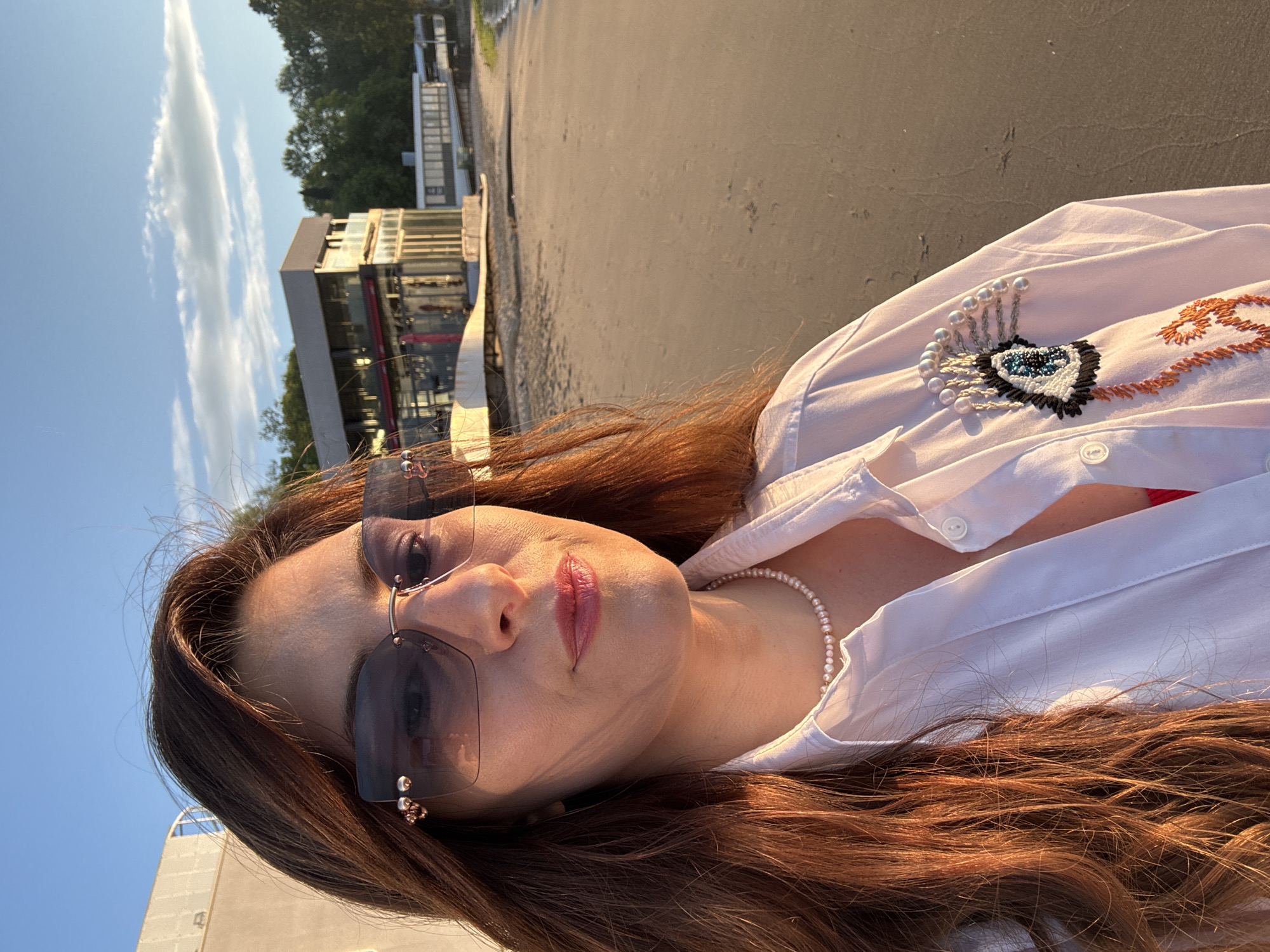 A medium shot of a woman wearing sunglasses, a white collared shirt with a beaded brooch in the shape of an eye, and a pearl necklace, taken on a beach near Varna, Bulgaria. The woman has long, reddish-brown hair. Behind her is a light tan sandy beach and buildings along the shoreline. The sky is a clear blue, with some wispy white clouds.