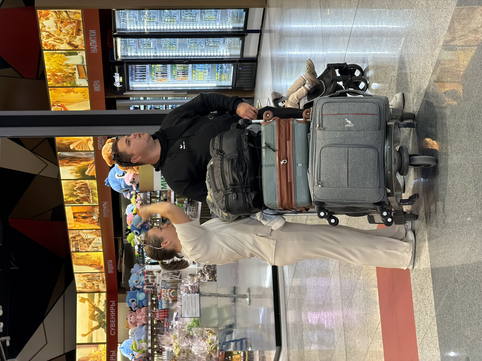 In the city of Khimki, Russia, at an airport retail area, a man and a woman stand beside a luggage cart laden with suitcases and backpacks. The man, wearing a black hoodie, has stuffed animals on his head. The woman, dressed in beige attire, is holding a stuffed toy. In the background, illuminated displays advertise souvenirs, food, and drinks, with refrigerated beverage coolers visible. A child is seated in a stroller attached to the luggage cart.