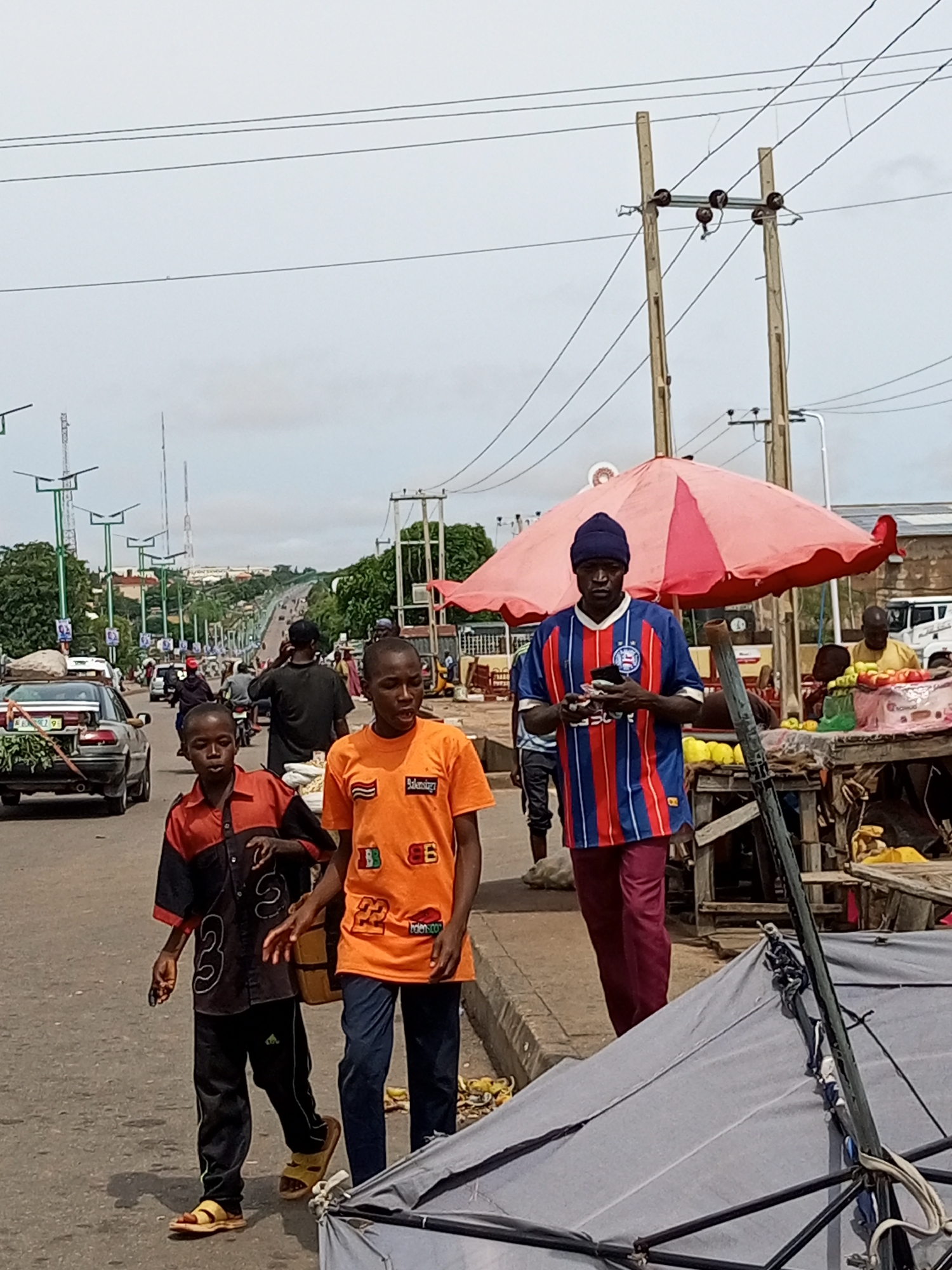 In Tumfure, Nigeria, a street scene unfolds under a cloudy sky, with a paved road stretching into the distance. Two young boys, one in an orange t-shirt and the other in a black and orange collared shirt with black pants and yellow sandals, walk towards the viewer on the right side of the road. To their right, a man wearing a blue, red, and white striped jersey and a dark blue beanie stands with a phone in his hand. Behind him, a stall with a pink umbrella displays fruit for sale, with other vendors and shoppers visible in the background. Vehicles, including a silver sedan with produce strapped to its roof and numerous motorcycles, navigate the road. Power lines crisscross the sky, supported by wooden poles.